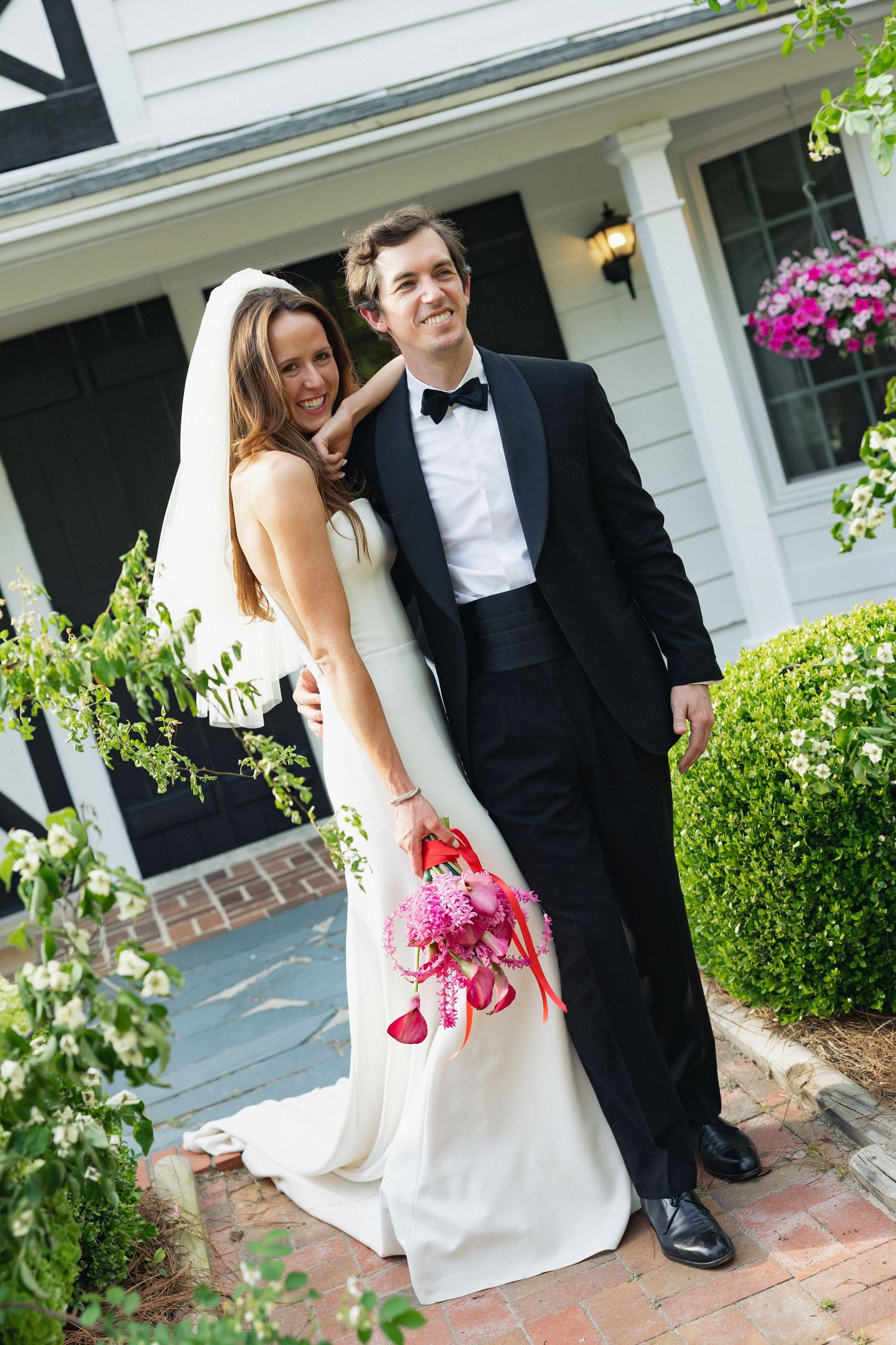 Bride in white gown and groom in black tuxedo pose together outside a charming house with pink bouquet.