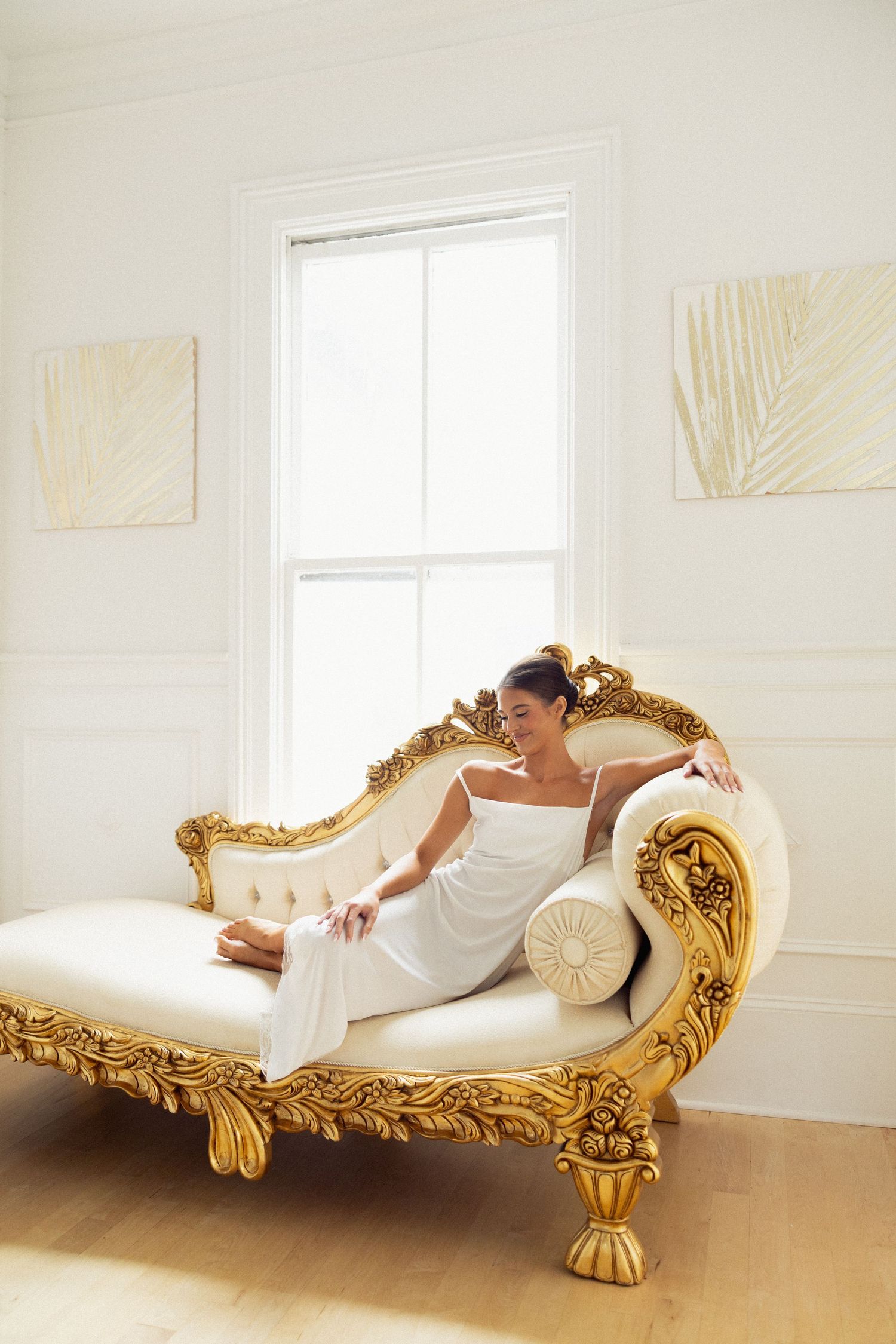 Woman in white dress reclining on ornate gold chaise lounge near bright window in elegant room.
