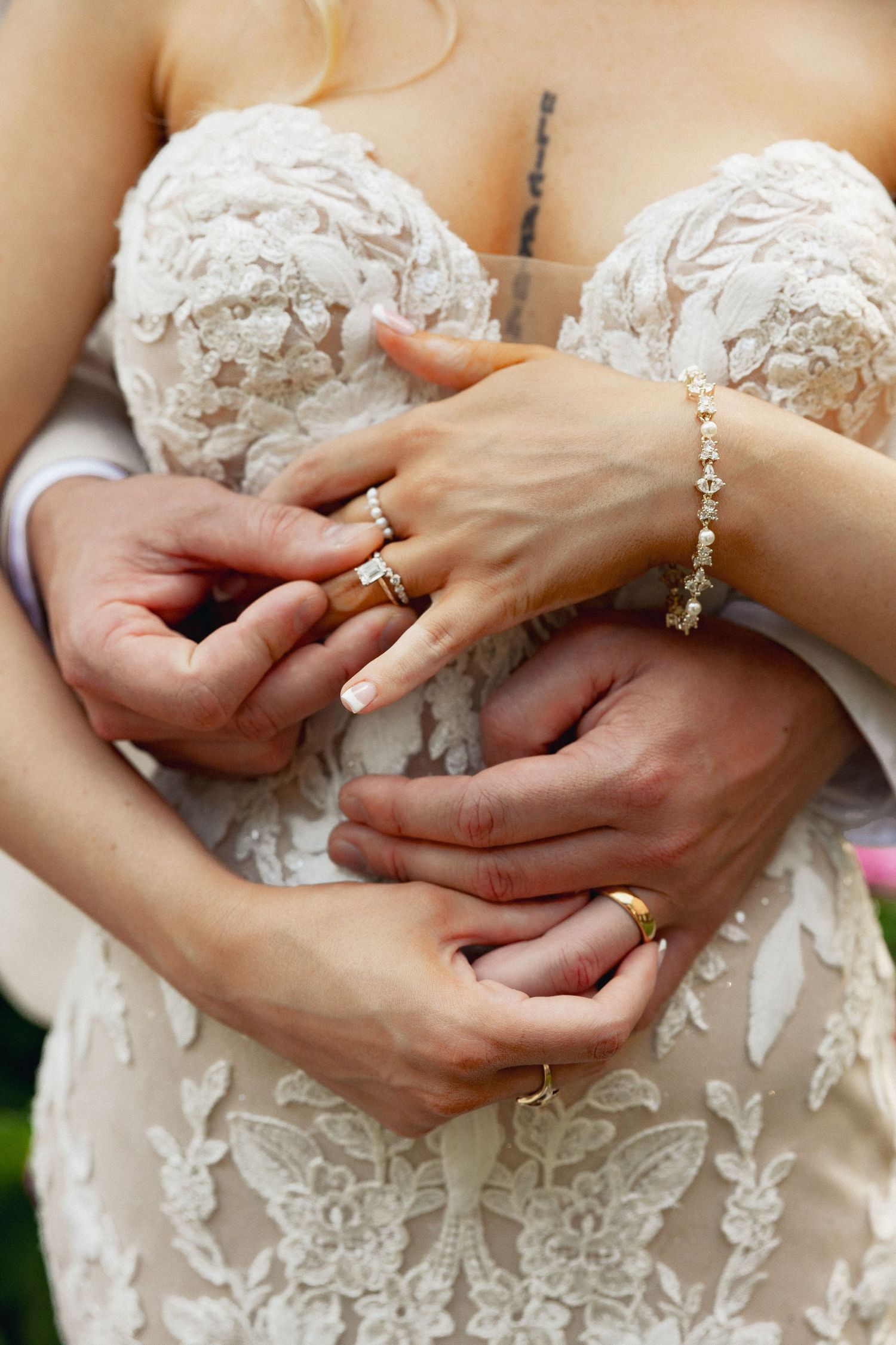 Bride and groom hands intertwined showing wedding rings and gold bracelet against lace wedding dress.