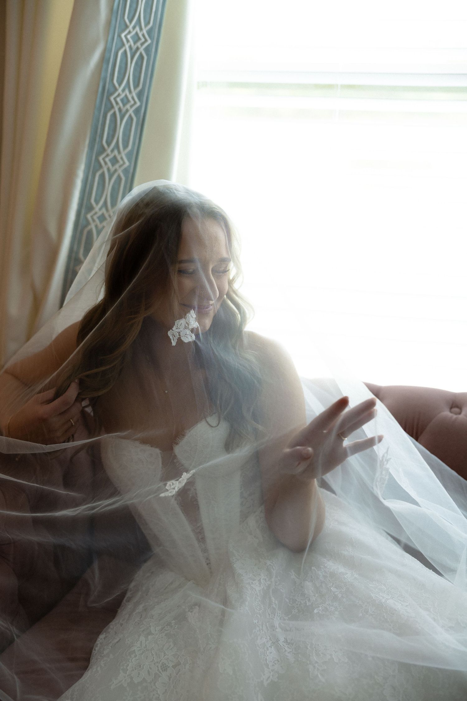 Bride in a lace wedding dress and veil sitting by a bright window, smiling softly in a dreamy bridal portrait.