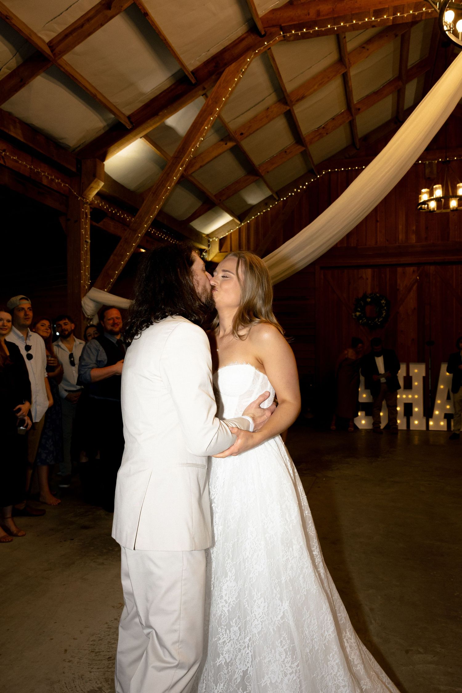 Bride and groom share first dance kiss in rustic barn wedding venue with string lights and white draping.