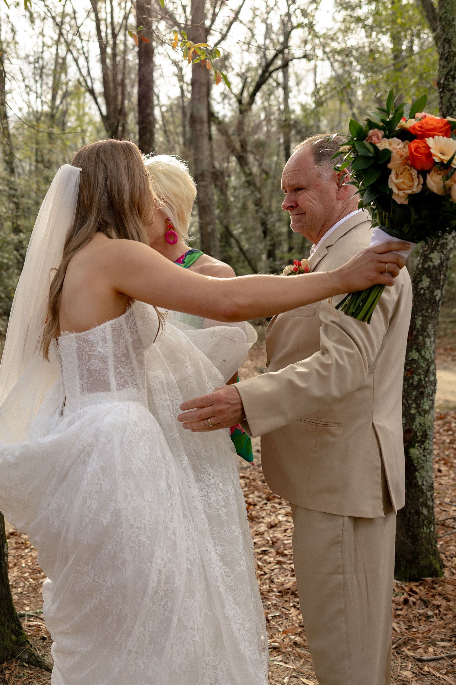 Bride in lace wedding gown greets man in tan suit during outdoor woodland wedding ceremony.