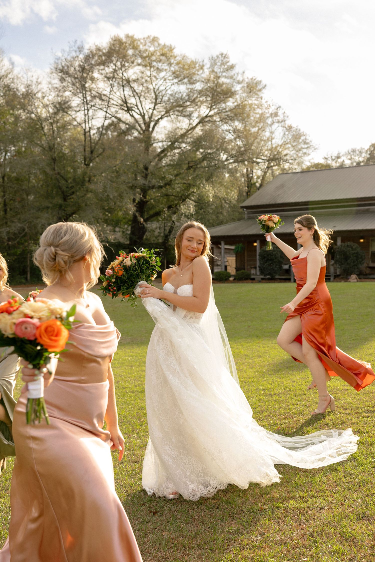 Smiling bride in white gown holding bouquet with bridesmaids in peach and rust satin dresses outdoors.