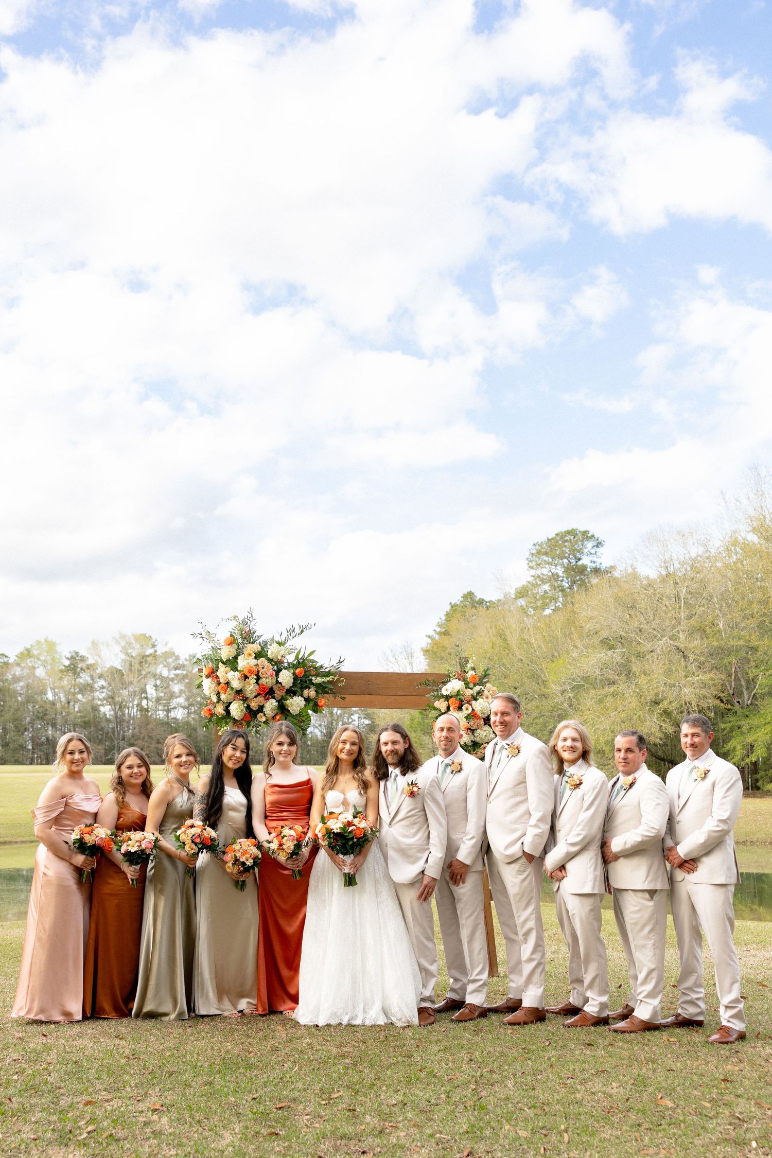 Wedding party in neutral and earth-tone attire poses under flower-adorned wooden arch.