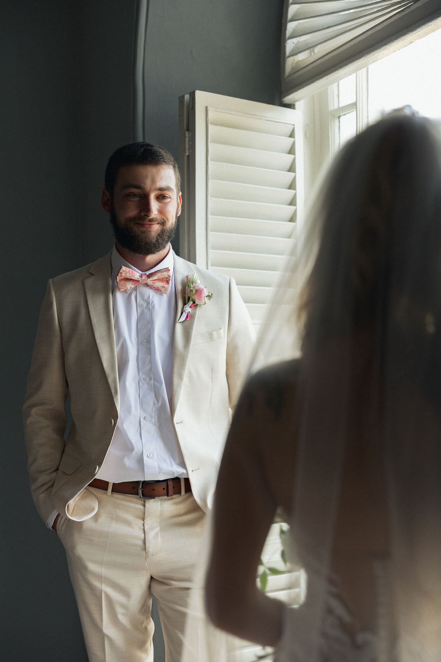 Bearded groom in beige suit with pink bow tie smiles at bride in veil near shuttered window.