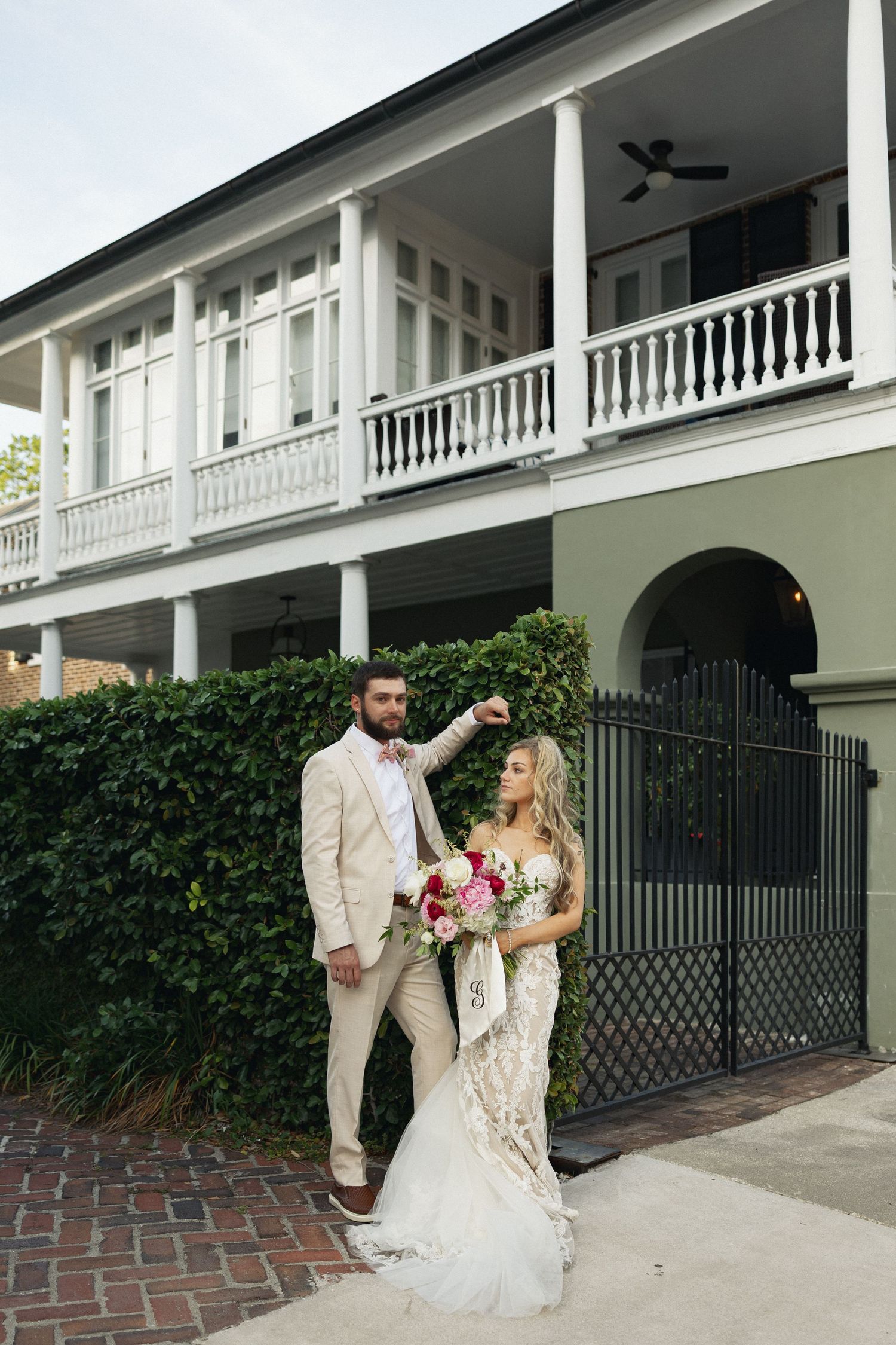 Bride and groom pose before historic Southern mansion with double porches and black iron gate.
