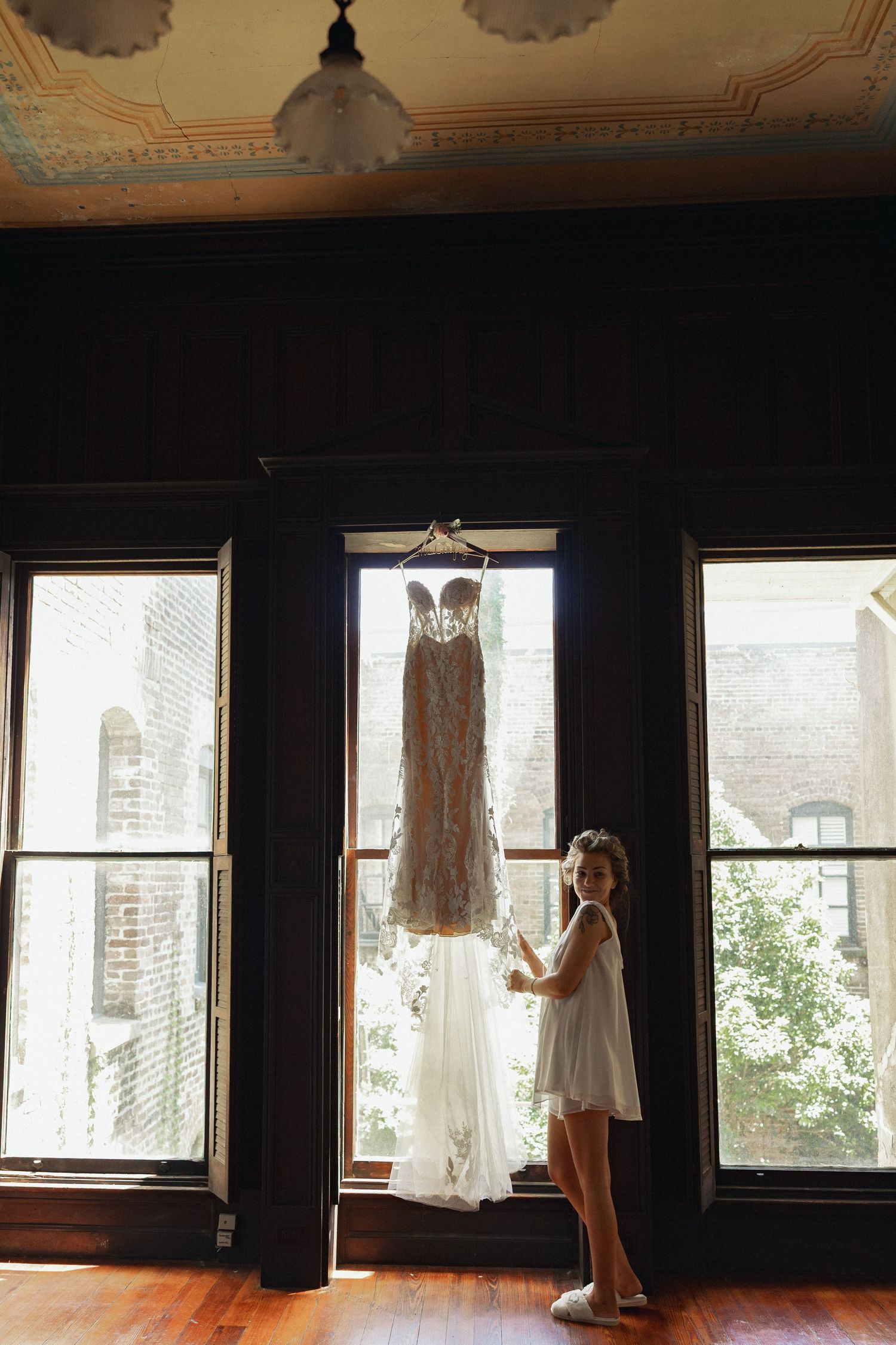 A flower girl in white dress admires a lace wedding gown hanging in a sunlit window of an elegant venue.