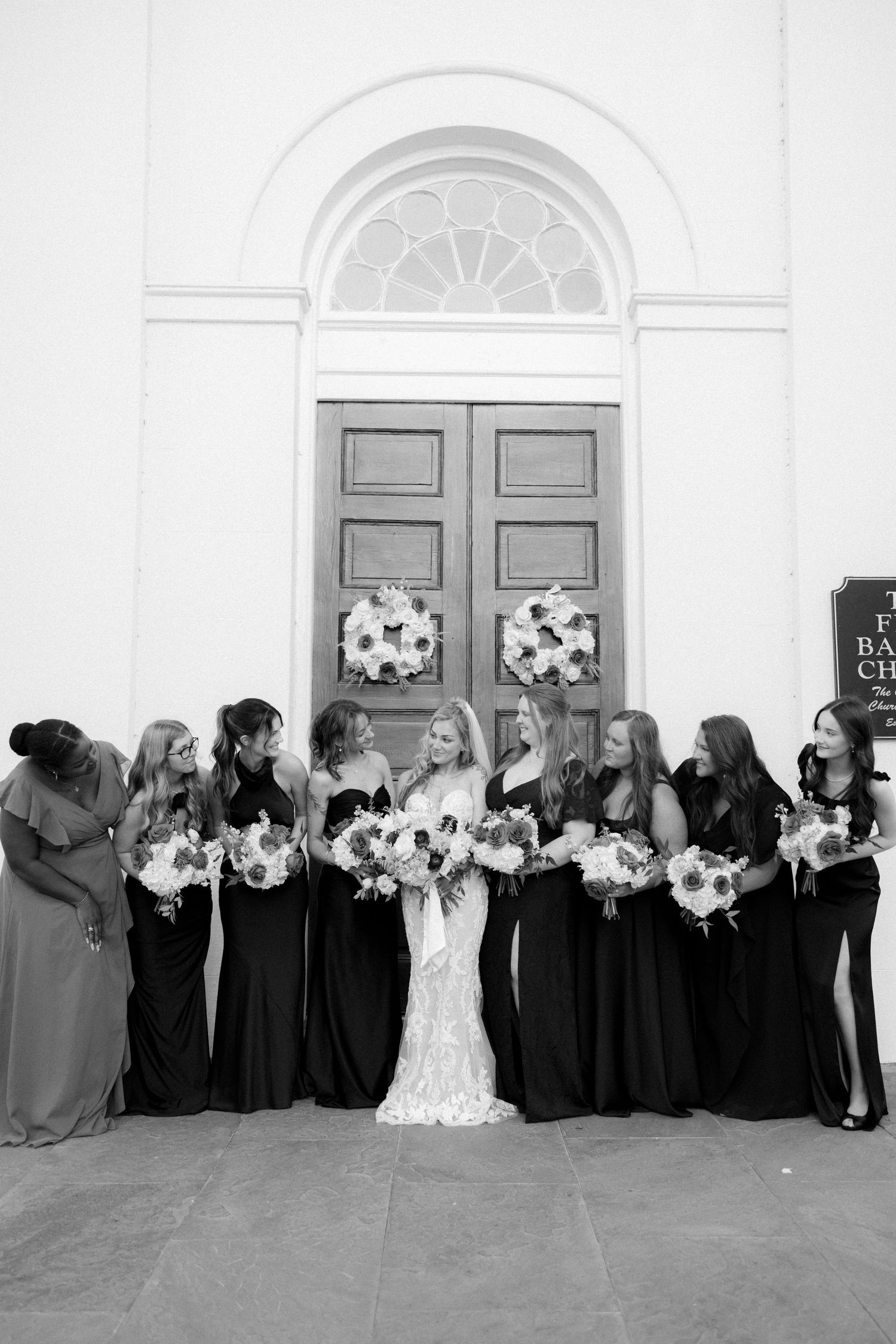 Bride poses with bridesmaids in black gowns holding floral bouquets outside a church.