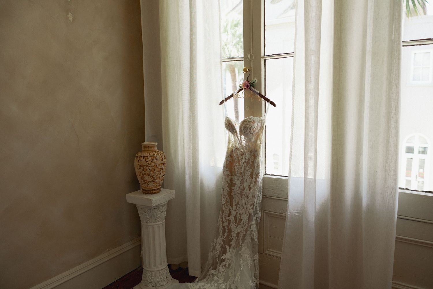 Elegant lace wedding dress hanging on wooden hanger in front of sheer white curtains by a bright window.