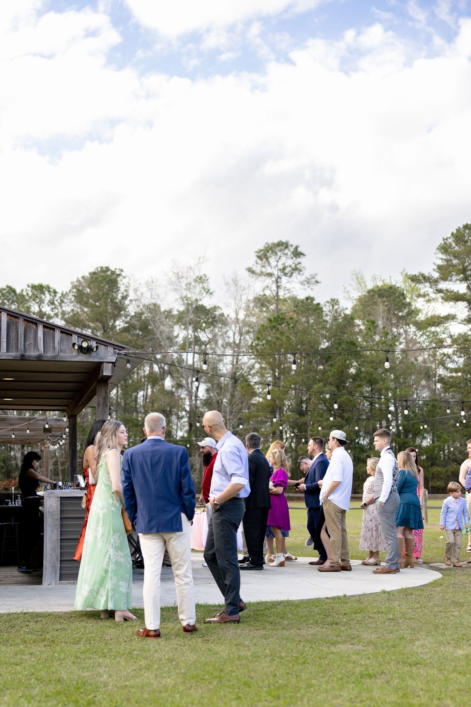 Wedding guests mingle outdoors on a patio near a bar area surrounded by lush green trees.
