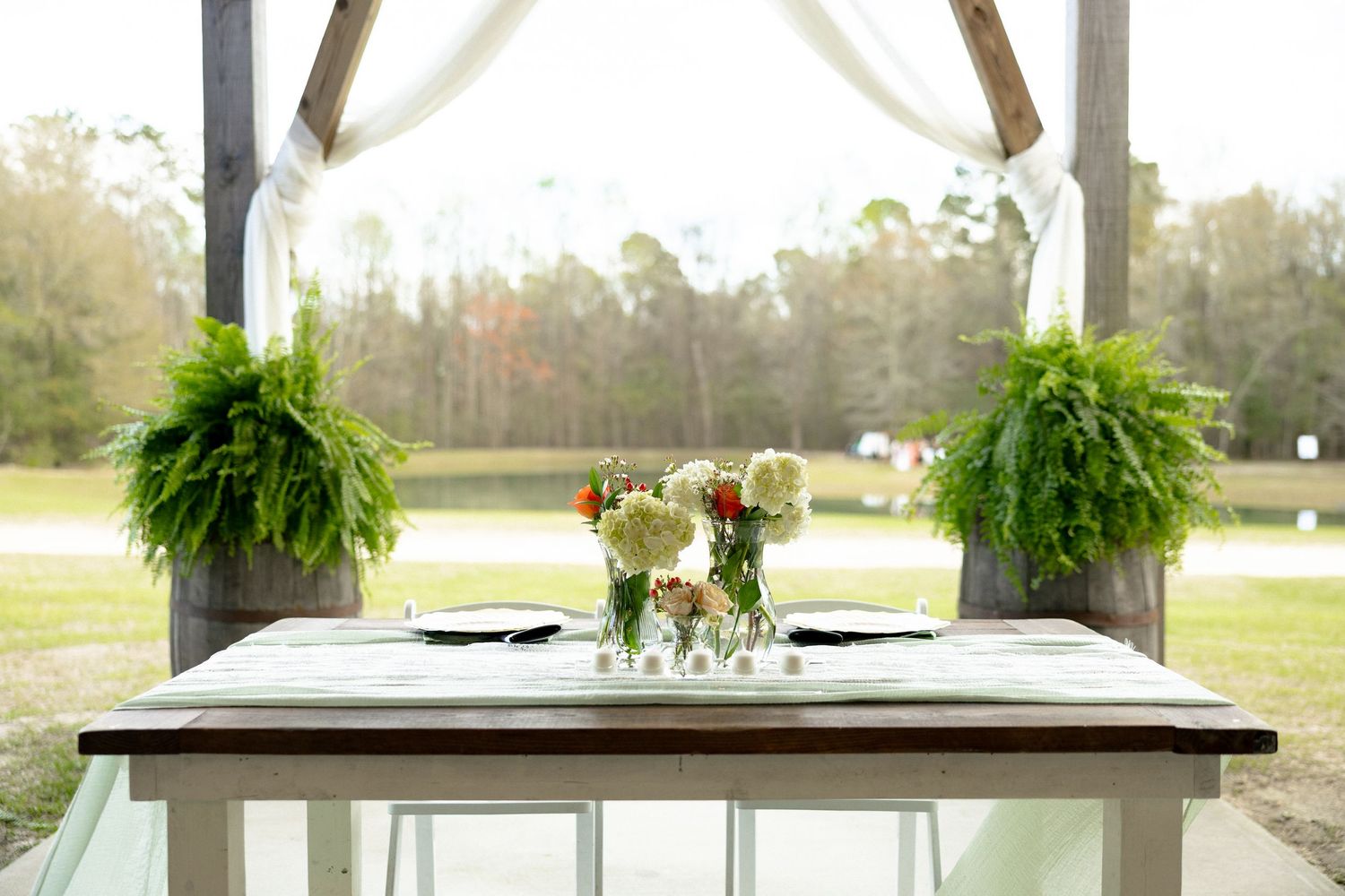 Sweetheart table with floral centerpiece and fern arrangements under a rustic barn pavilion.