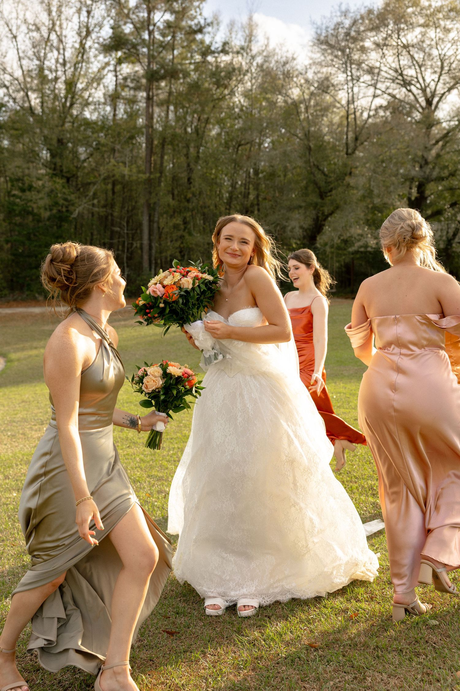 Bride in white lace gown smiling at camera surrounded by bridesmaids in mixed earth-tone dresses outdoors.