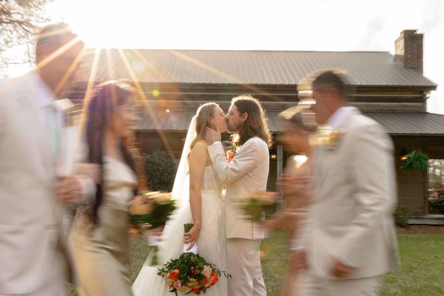 Newlyweds share a kiss in golden sunlight as wedding party moves around them.
