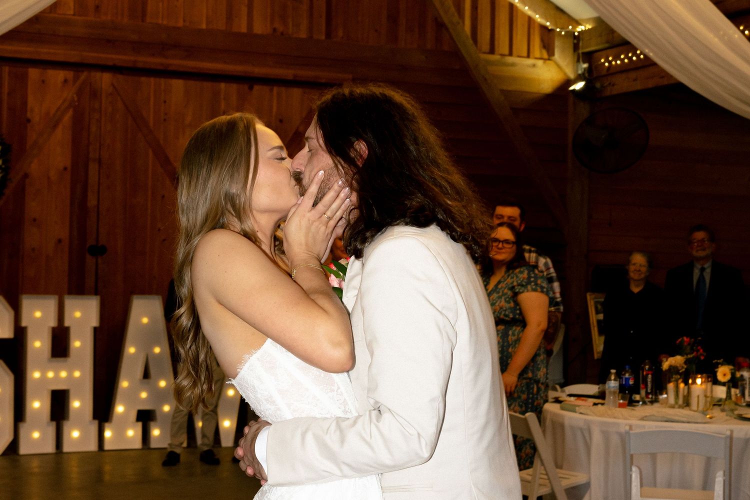 Bride and groom share a passionate kiss in front of illuminated SHAW marquee letters at their wedding.