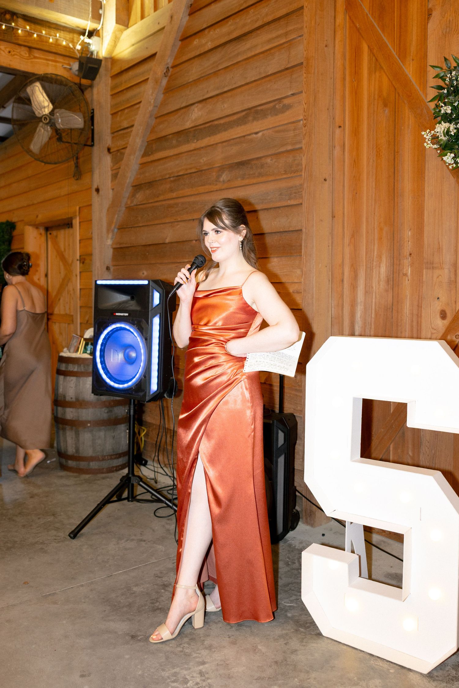 Woman in rust orange satin gown holds microphone next to speaker at rustic barn wedding reception.