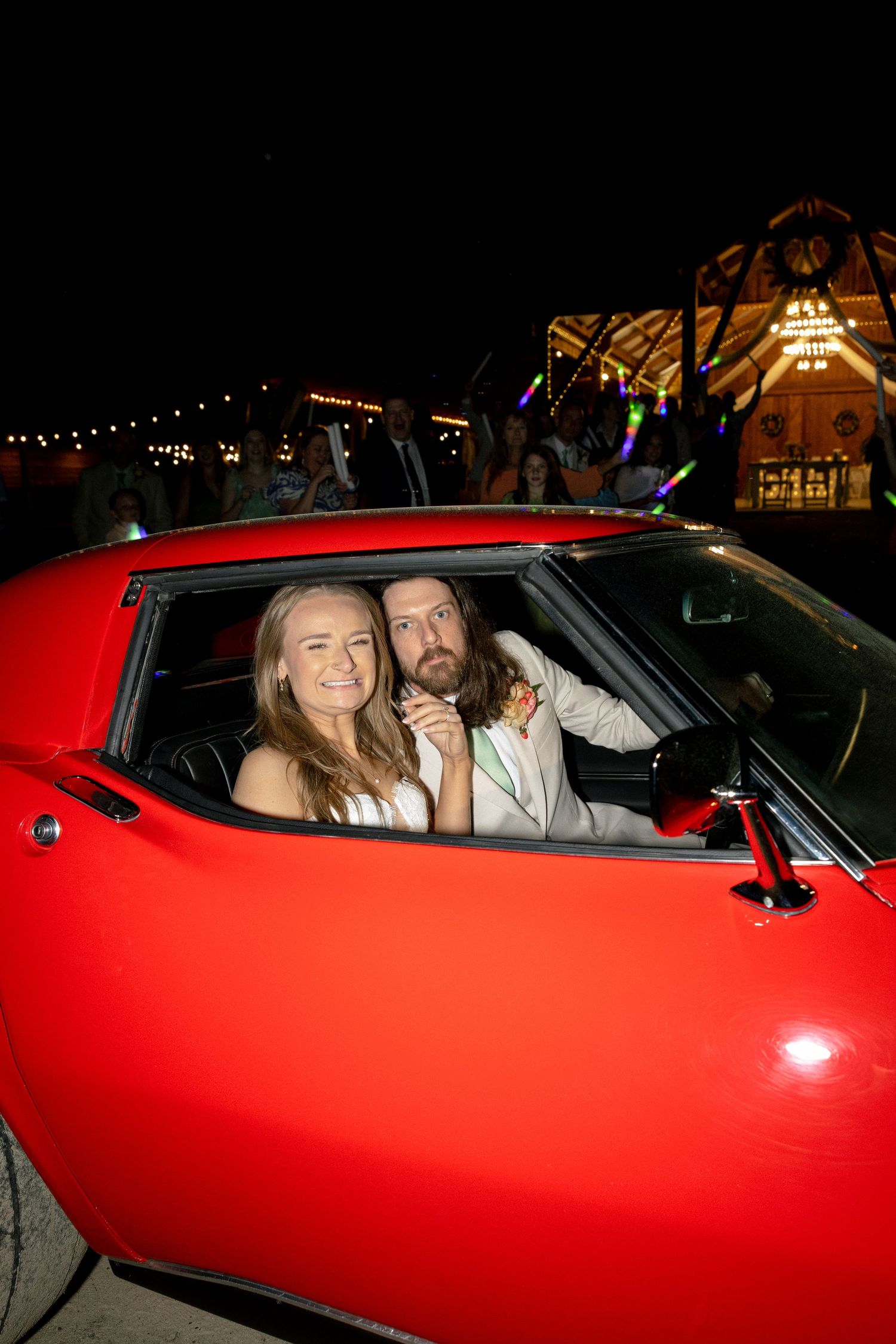 Couple smiling in a classic red sports car at night with festive lights in the background.