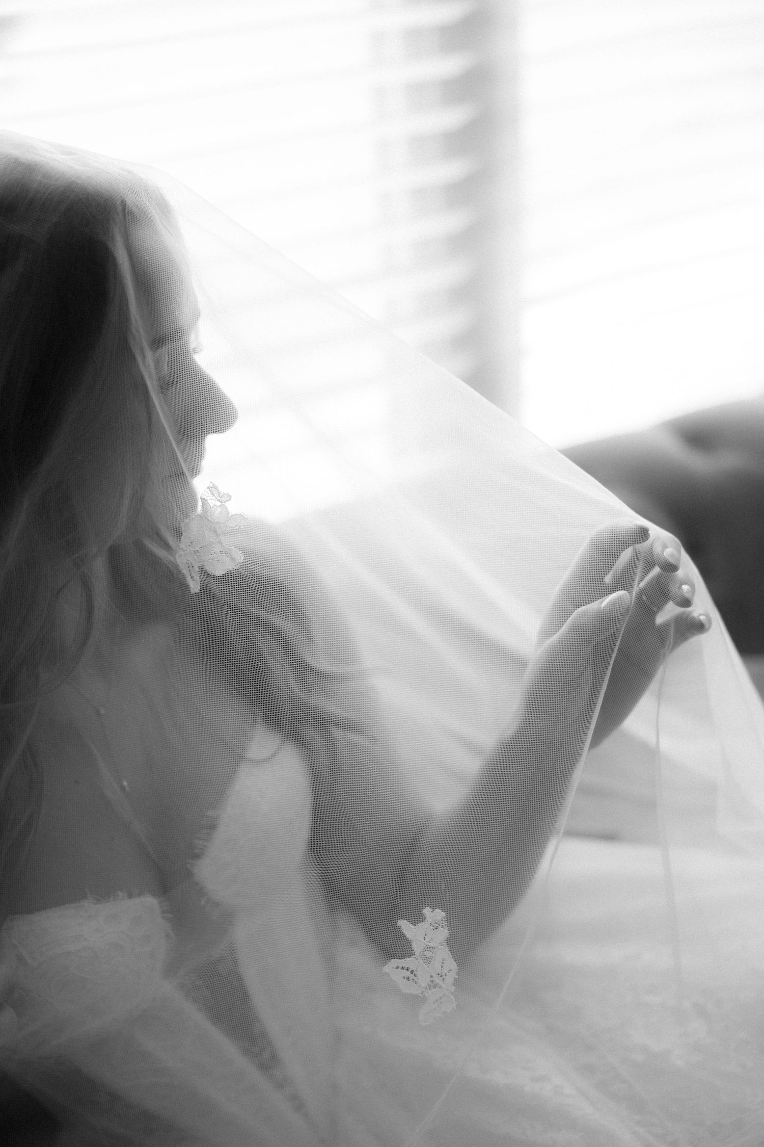 Black and white portrait of bride draped in floral veil, soft light streaming through window.