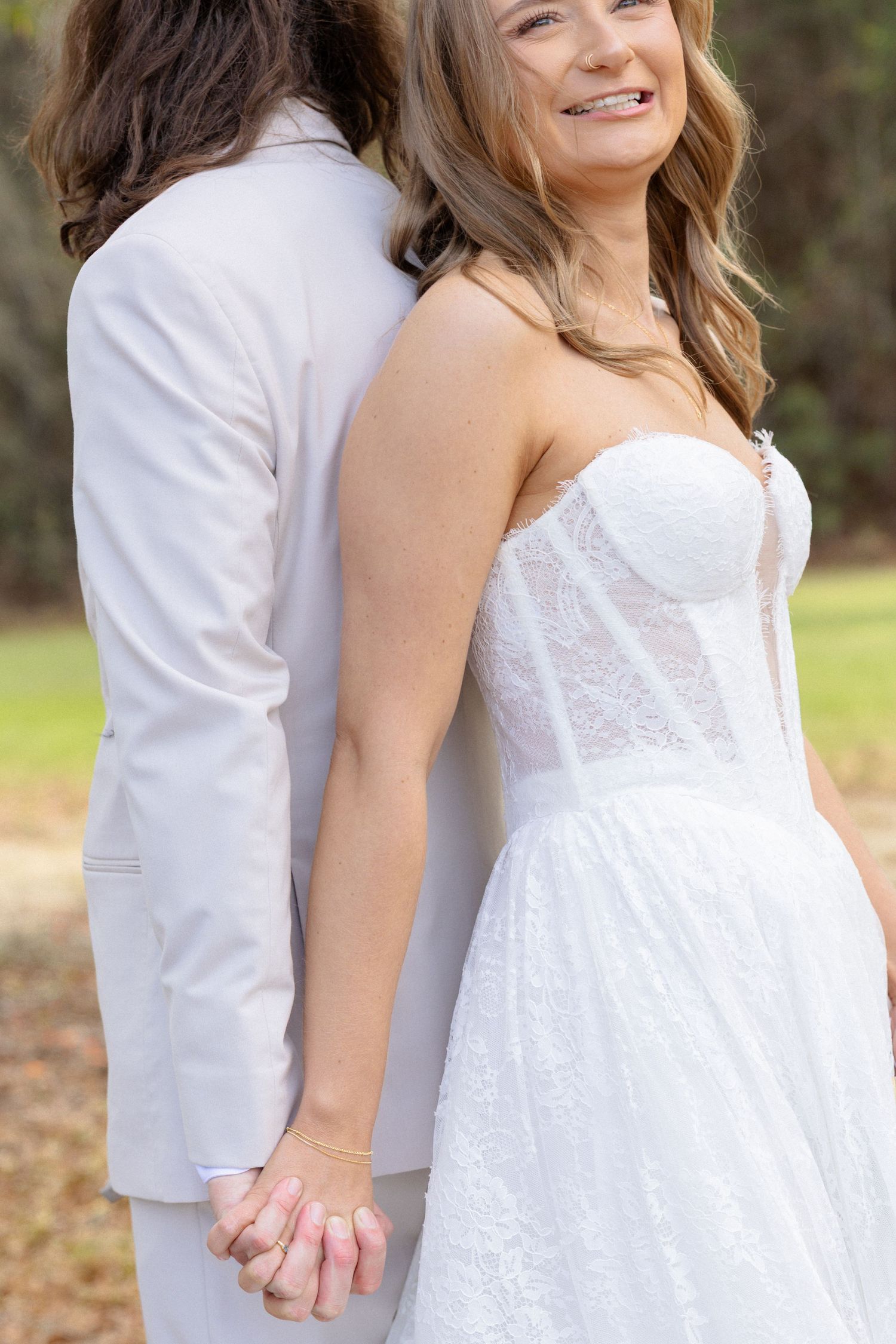 Smiling bride in strapless lace corset wedding gown holds hands with groom in beige suit outdoors.