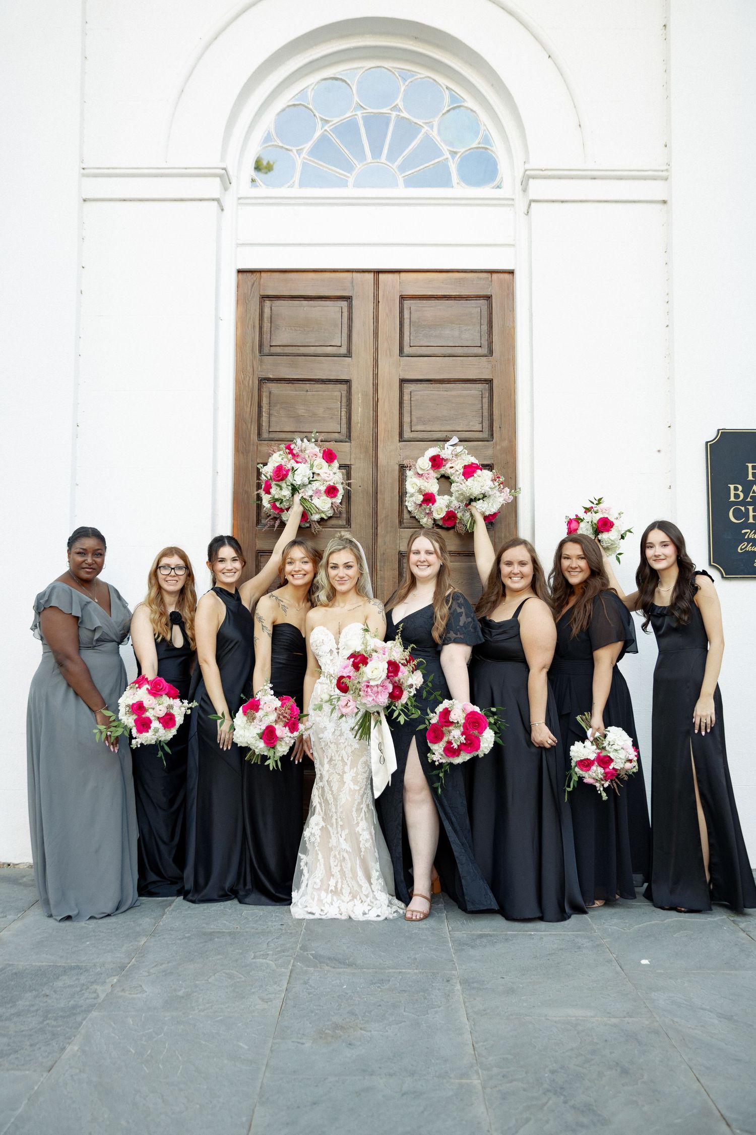 Bride in lace gown poses with bridesmaids in black dresses holding red bouquets outside white church.