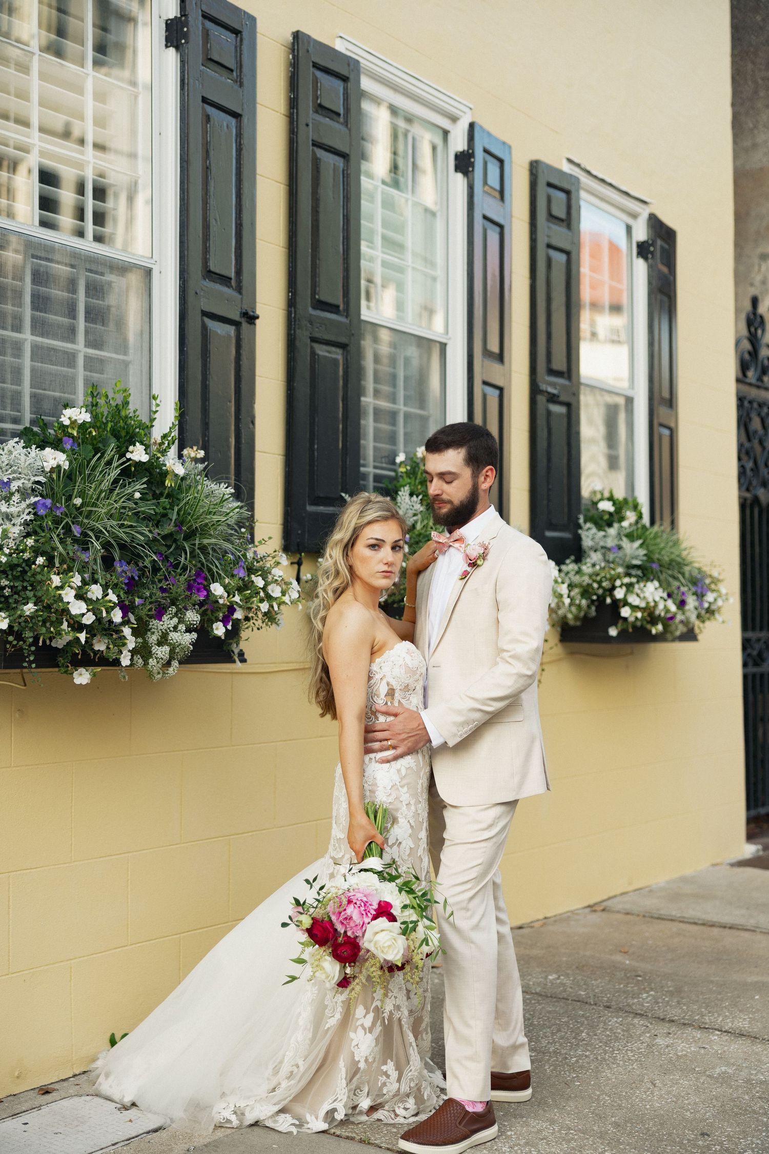 Wedding couple embracing on Charleston street, bride in lace gown holding pink bouquet, groom in tan suit, yellow building.