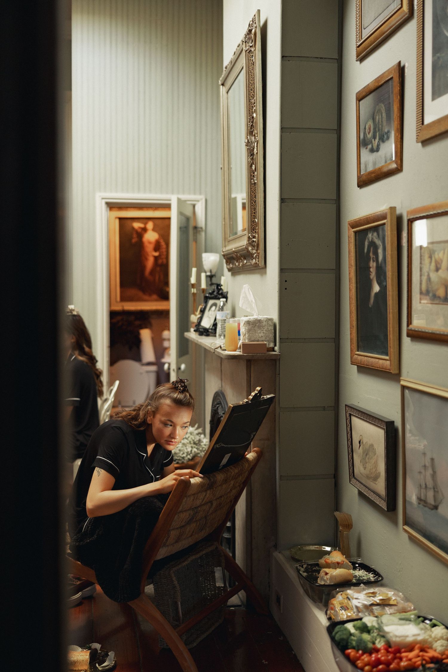 Artist painting at an easel in a narrow hallway lined with framed artwork and vintage portraits.