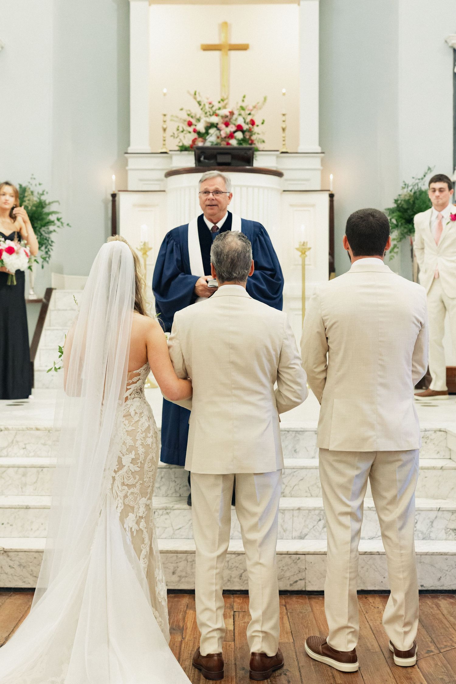 Bride and groom stand before officiant at church altar during traditional wedding ceremony with cross backdrop.