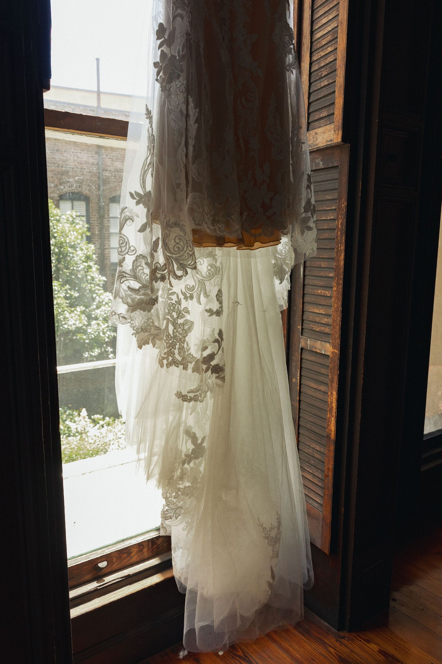 Lace wedding dress hanging in a sunlit window with wooden shutters.
