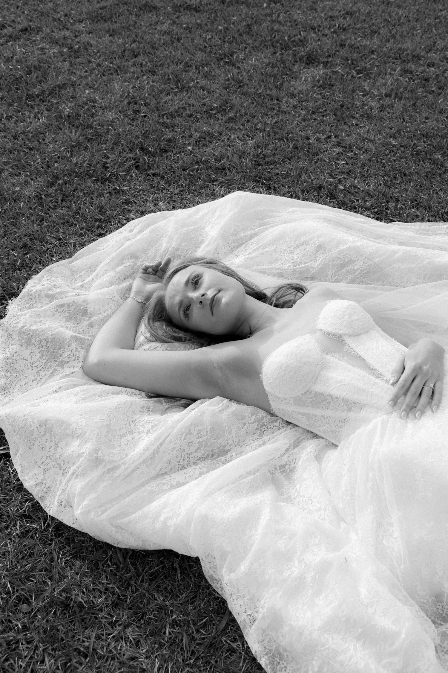 Black and white photo of a bride in a white gown lying on grass, with her dress spread around her.