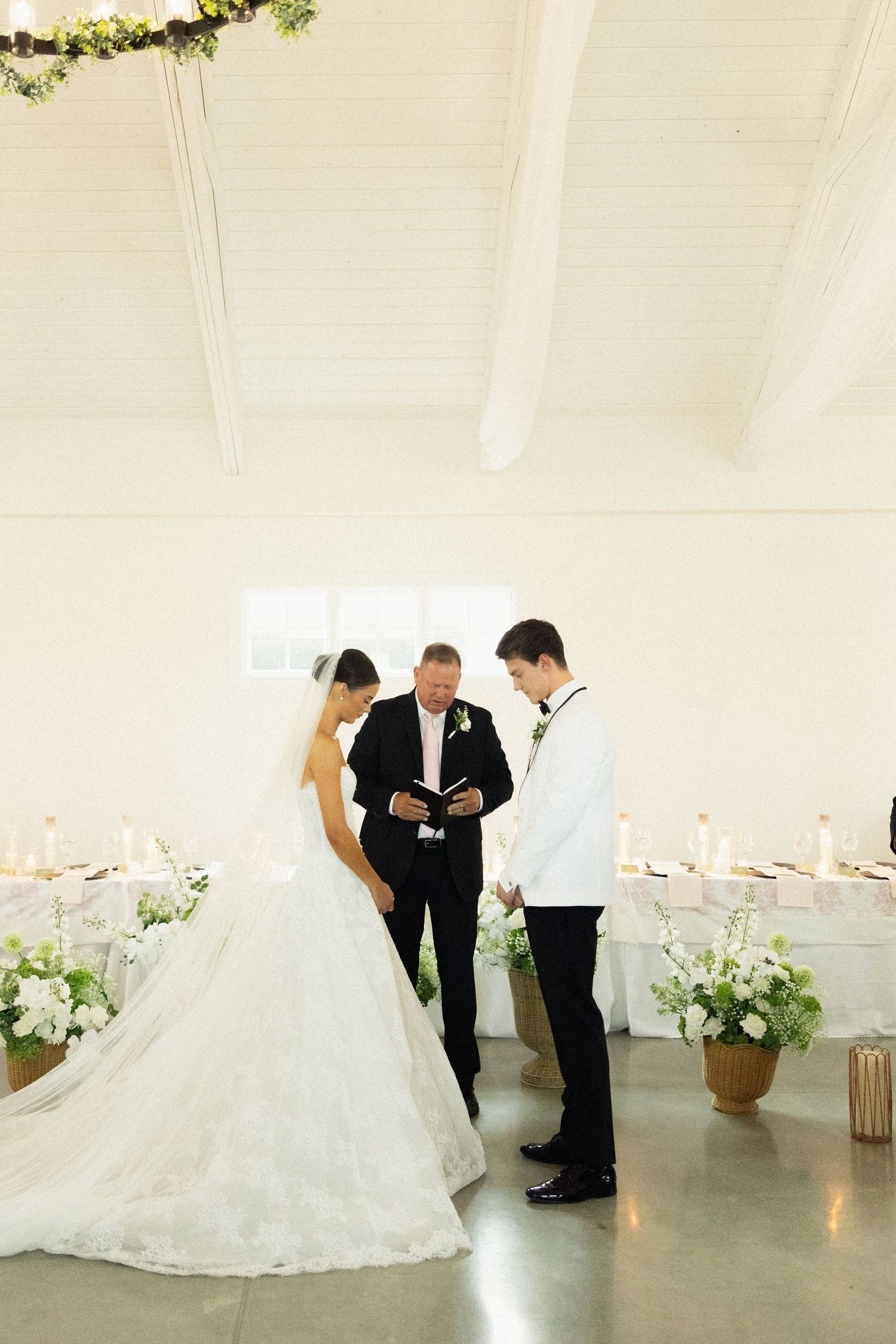 Bride and groom exchanging vows with officiant in elegant white venue adorned with floral arrangements.