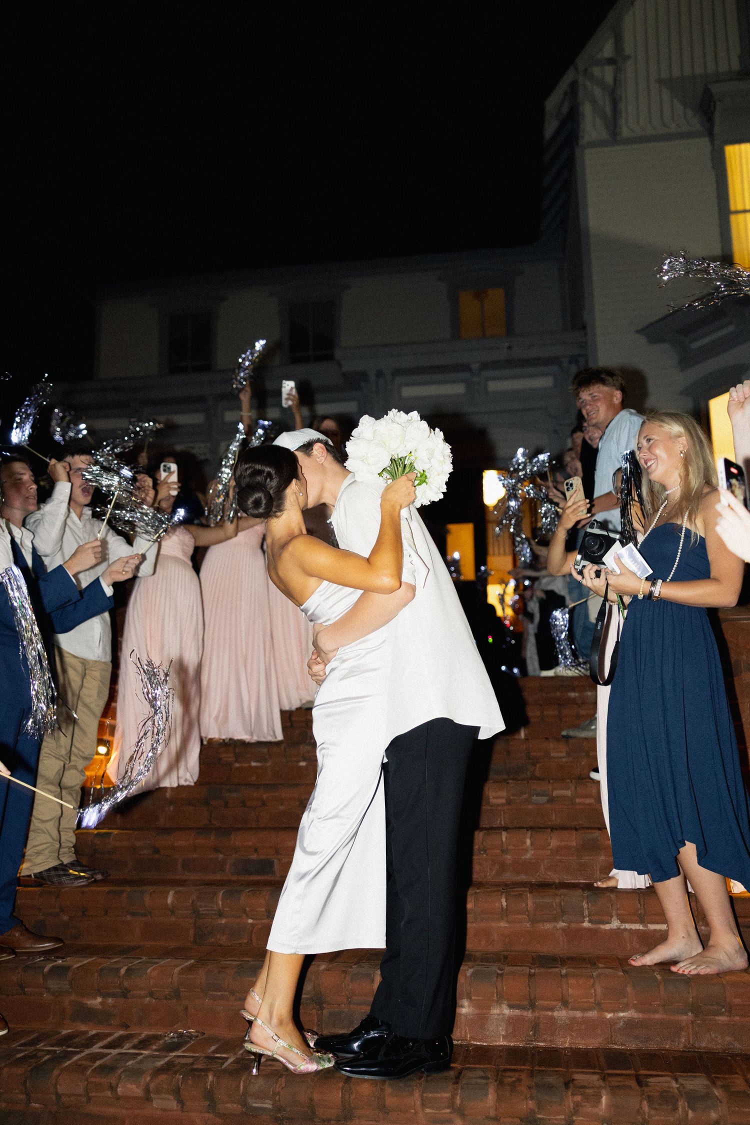 Bride and groom share a romantic kiss on brick steps surrounded by cheering wedding guests at night.