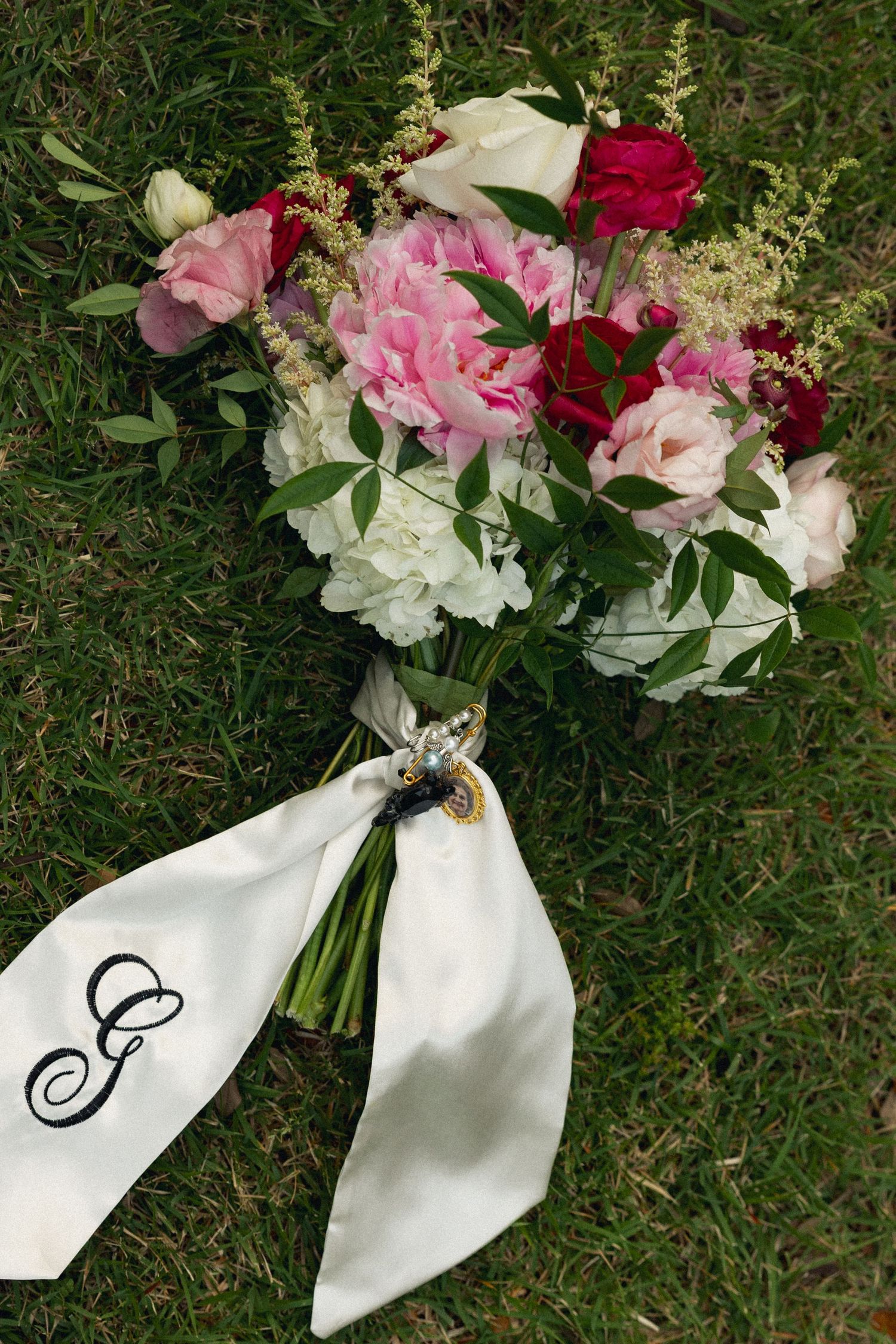 Bridal bouquet with pink peonies, red roses, and white flowers tied with monogrammed satin ribbon on green grass.
