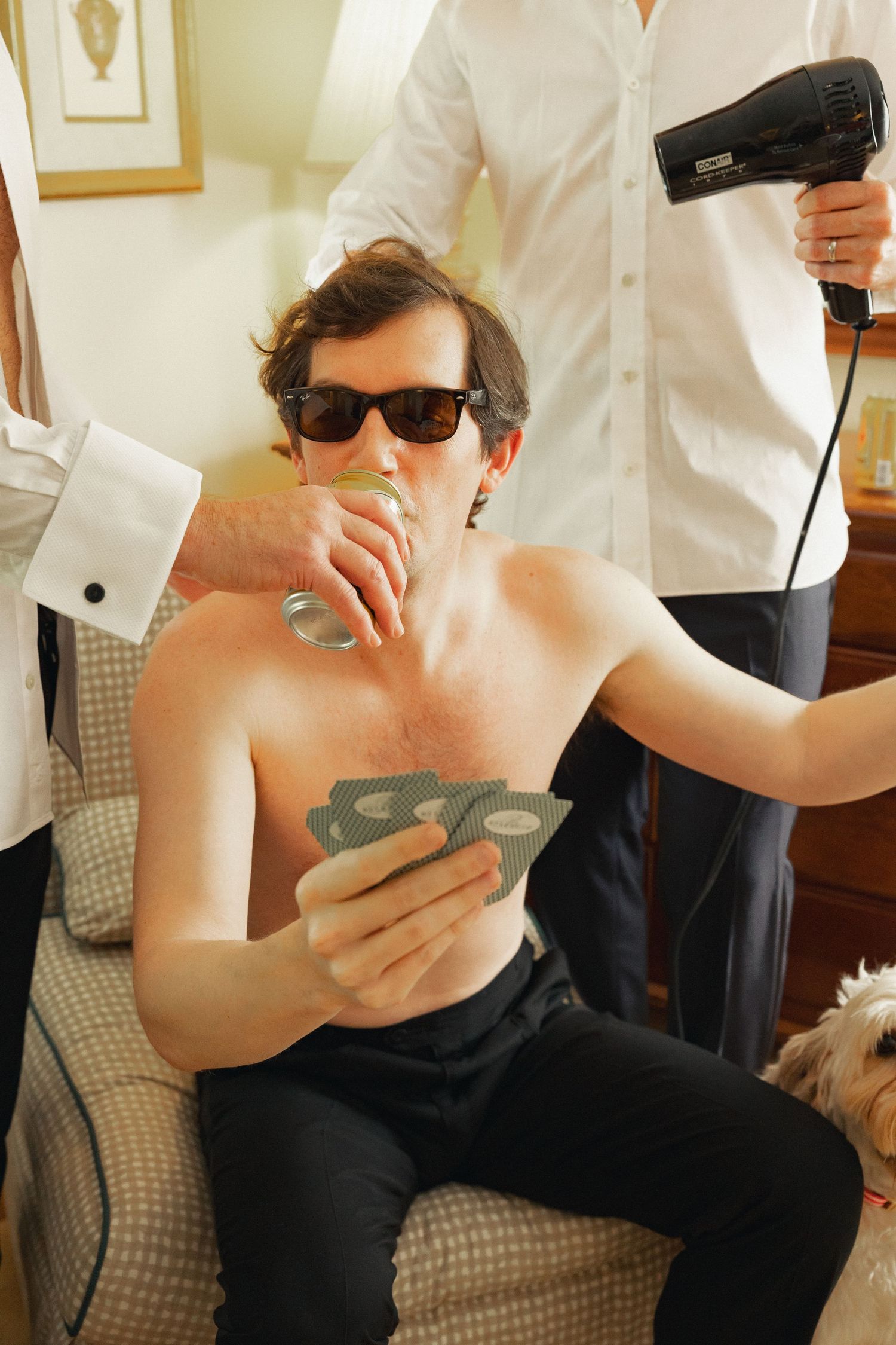 Shirtless man in sunglasses holds cards and drinks while someone blow-dries his hair in a hotel room.