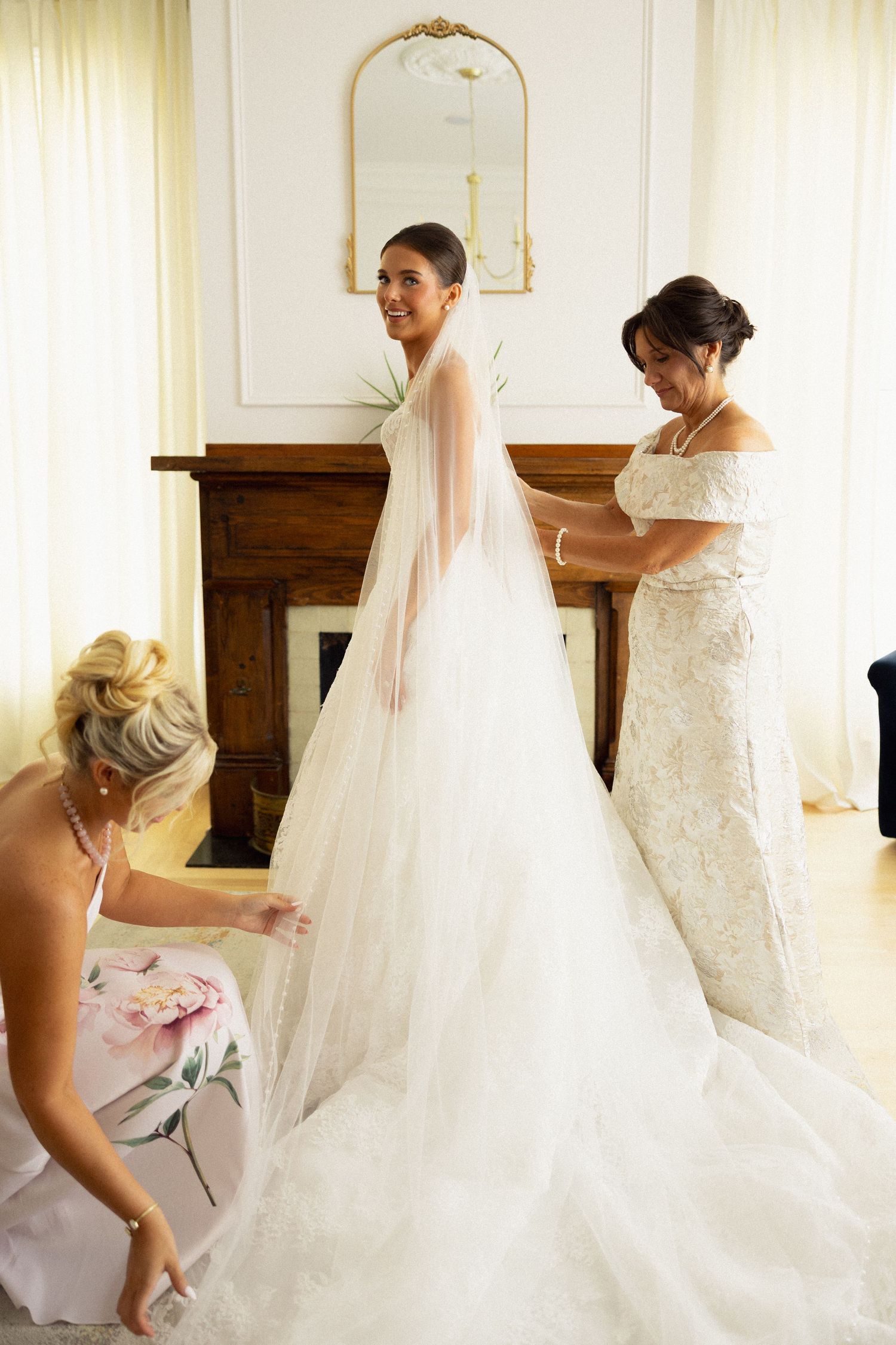 Bride in white ball gown with veil being helped by bridesmaid and mother before wedding ceremony.