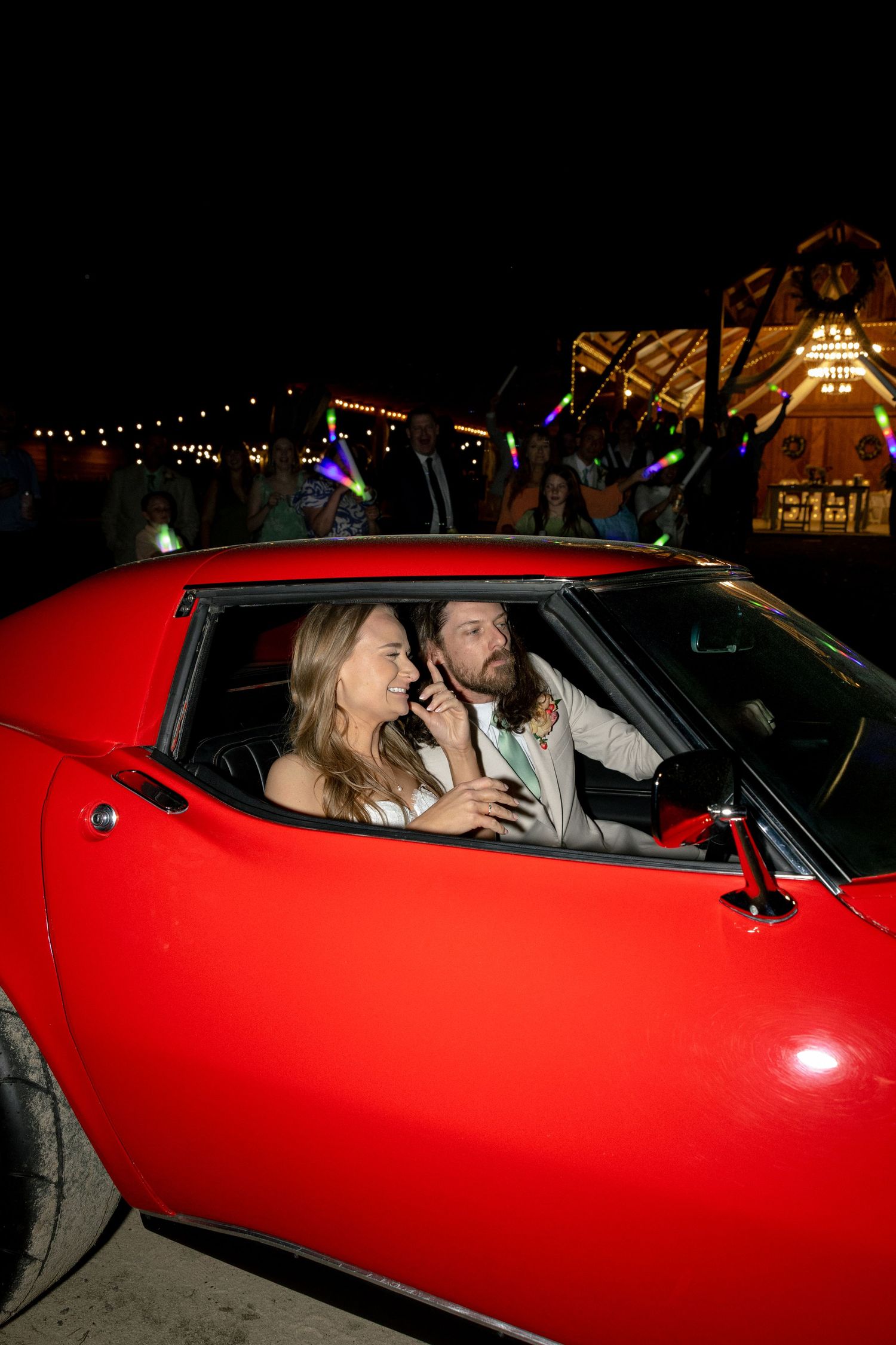 Smiling newlyweds ride together in a red classic sports car during their nighttime wedding departure.