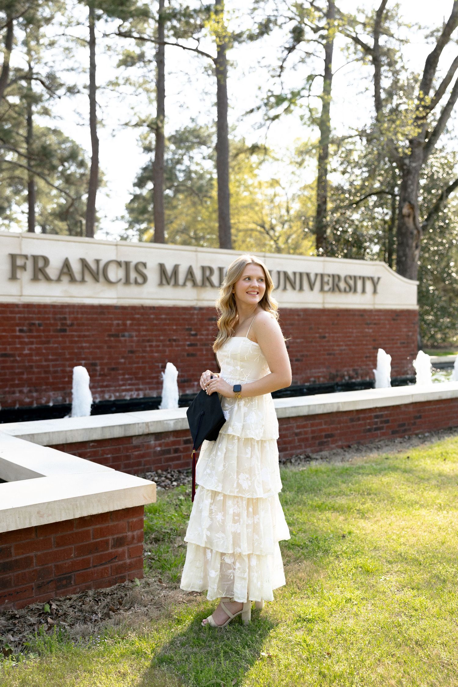 Smiling graduate in white dress posing in front of Francis Marion University sign.