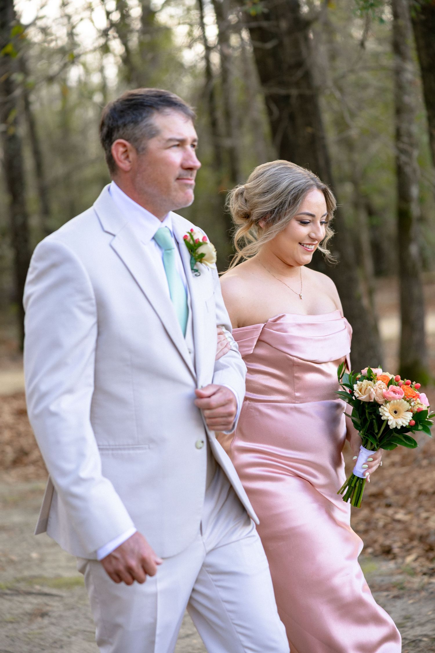 A groomsman in white suit with mint tie walks alongside a bridesmaid in blush pink off-shoulder gown at outdoor wedding.