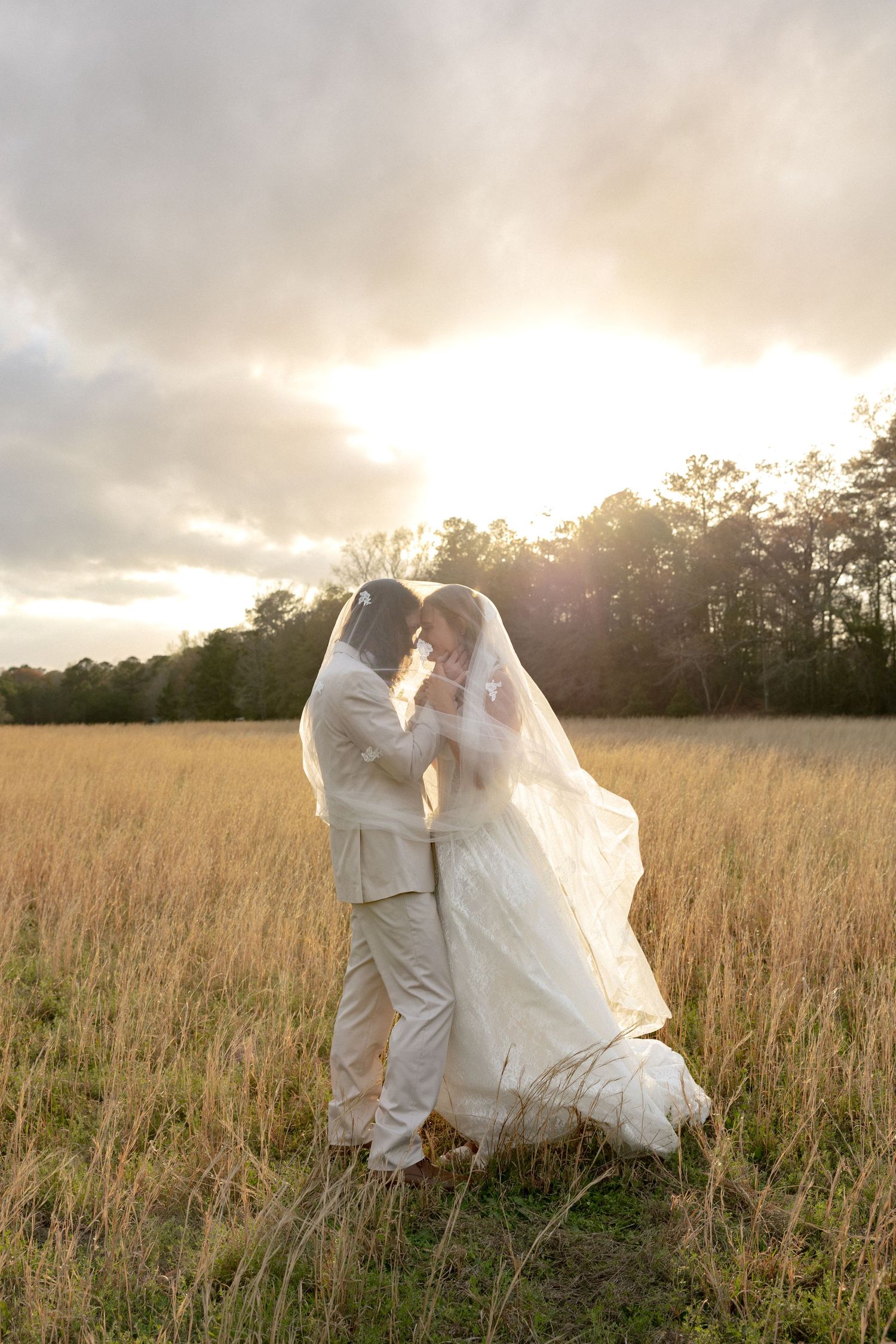 Newlyweds kiss under billowing veil in golden field with stunning sunset backdrop.