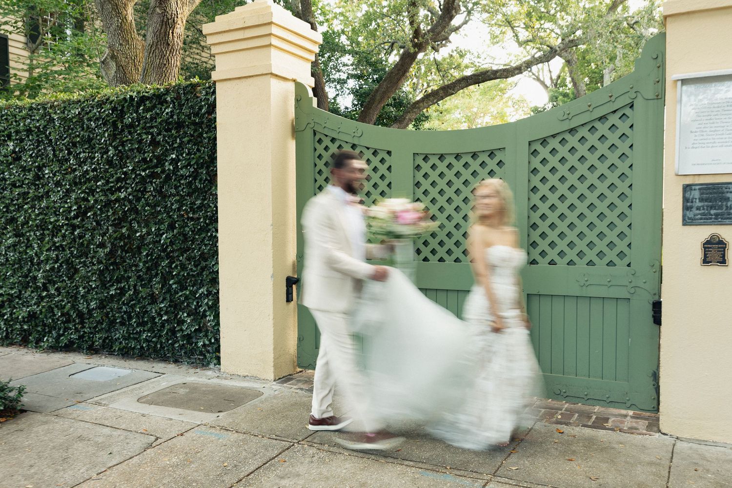 Bride and groom walk through green lattice garden gate in motion-blurred wedding photo.