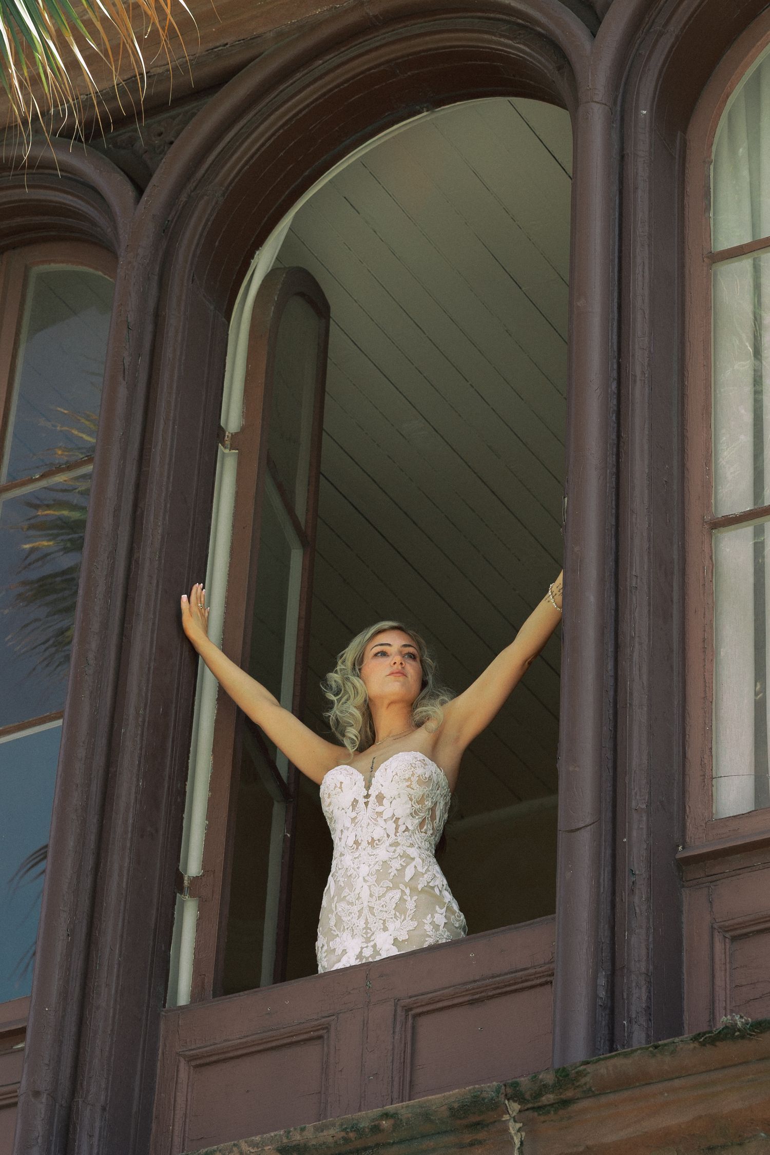 Bride in lace wedding gown stands in arched window with arms raised at historic building.