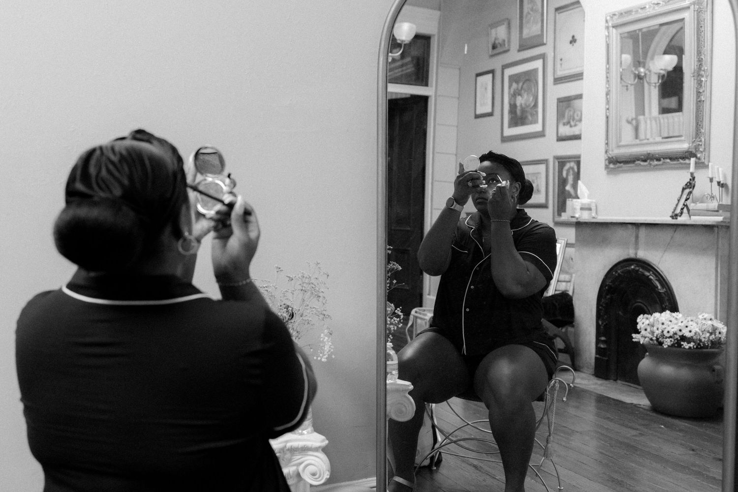 Black and white photo of a woman photographing herself in a mirror, surrounded by framed artwork and a fireplace.