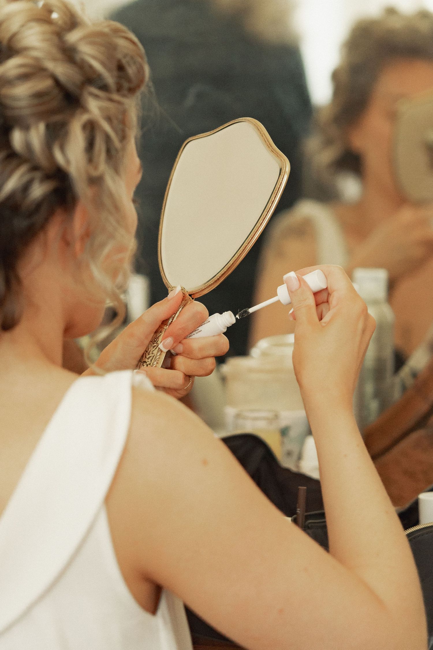 A bride holds a decorative hand mirror while applying mascara during her wedding day preparations.