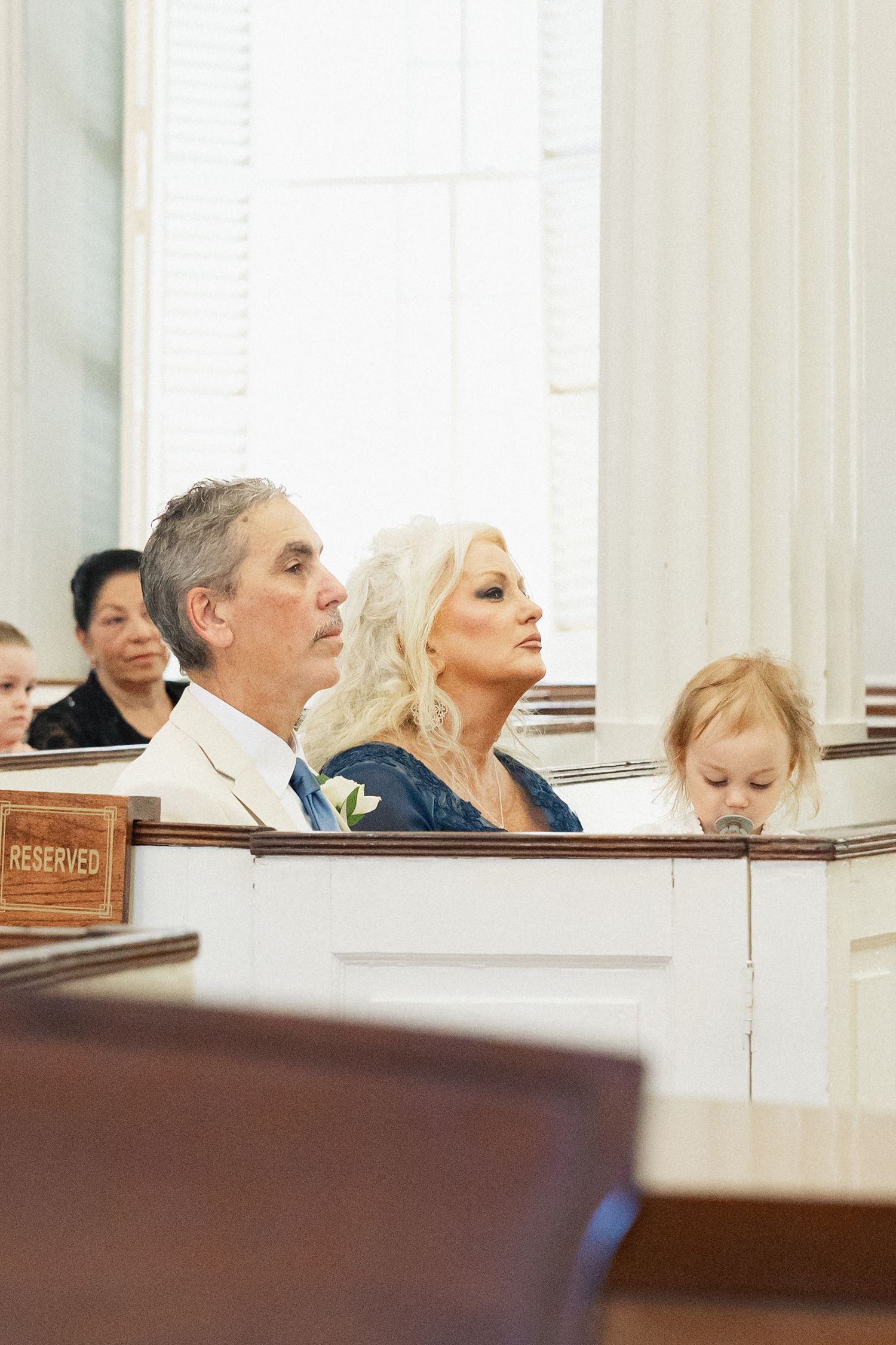 Wedding guests including a couple and small child seated in reserved church pews during ceremony.