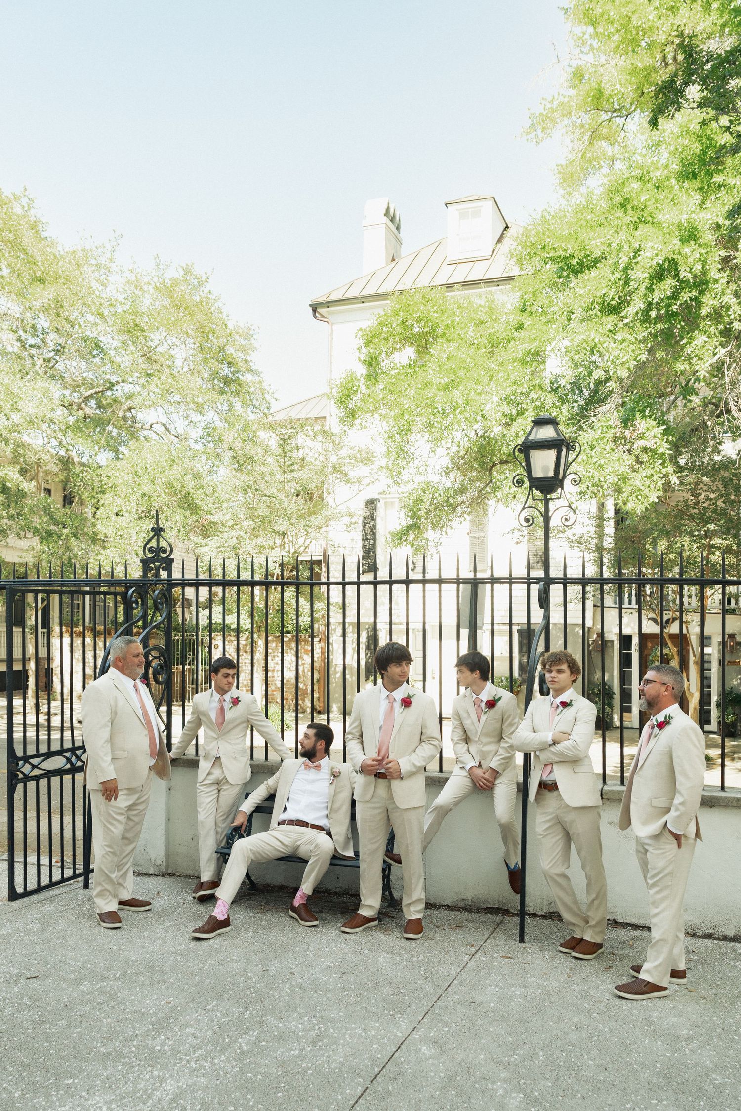Groom and groomsmen in cream suits lean against iron fence outdoors on sunny wedding day.