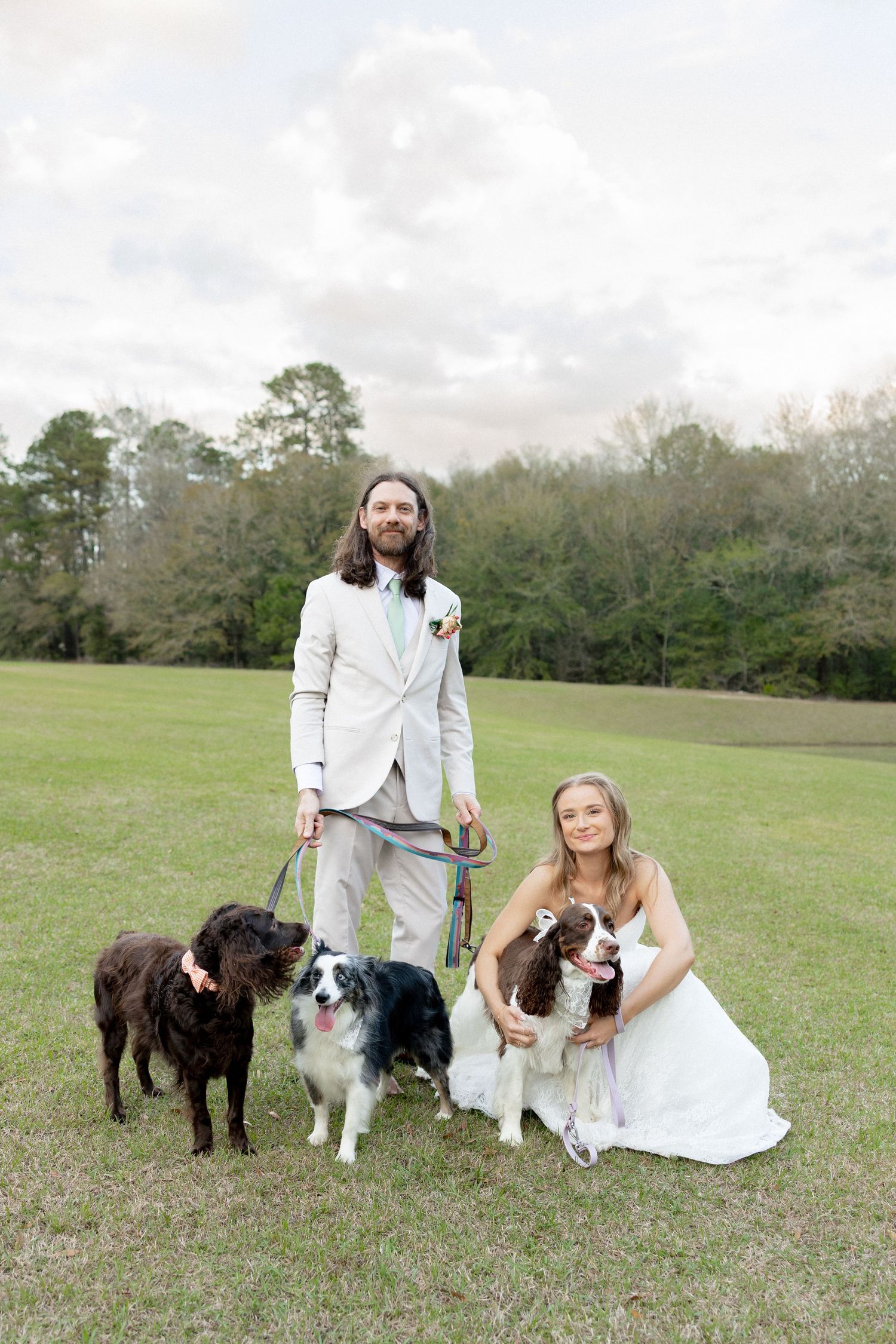 Bride and groom pose with three dogs on green lawn in wedding attire, trees and cloudy sky in background.