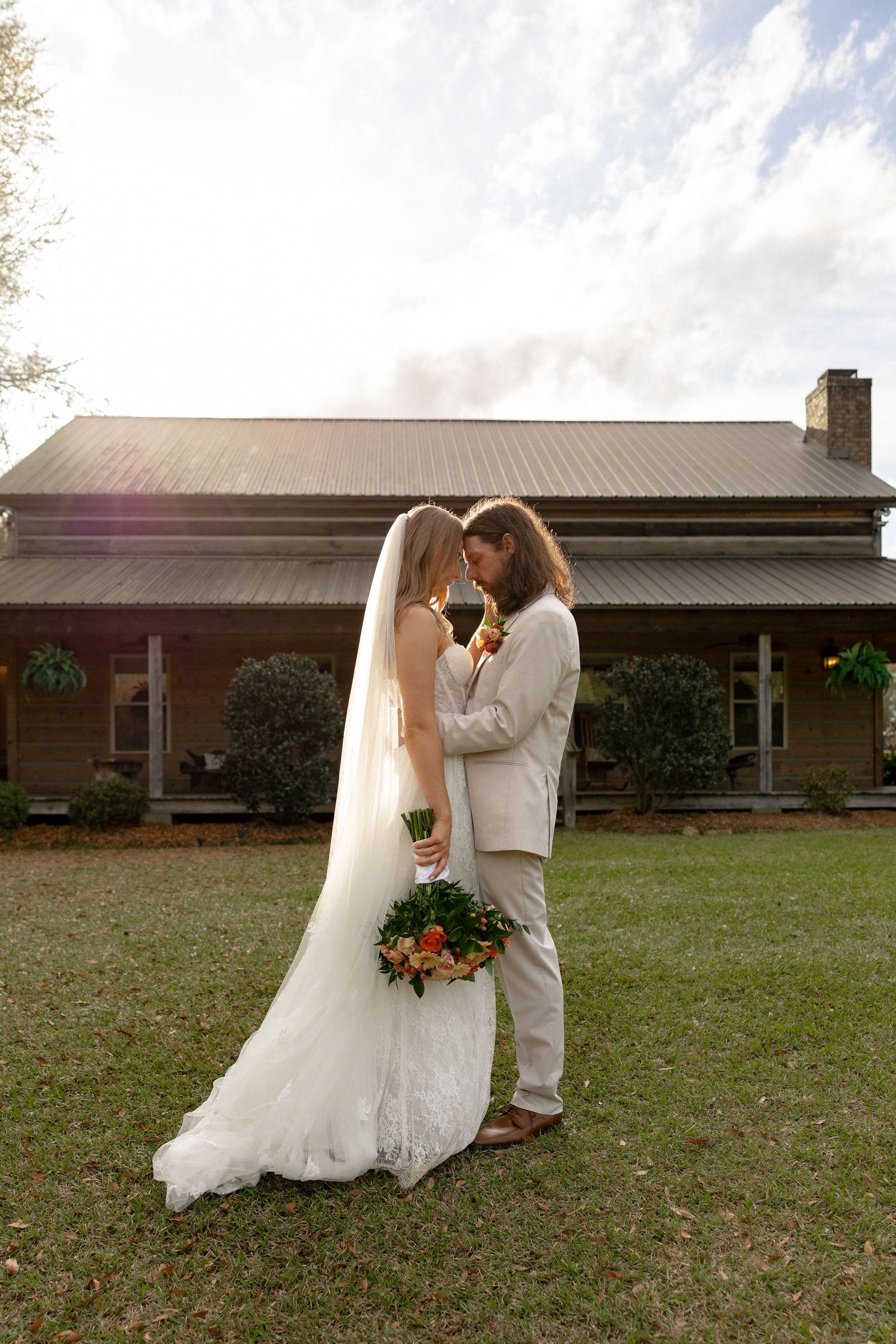 Bride and groom sharing an intimate kiss in front of a rustic farmhouse, bride in lace gown with long veil.
