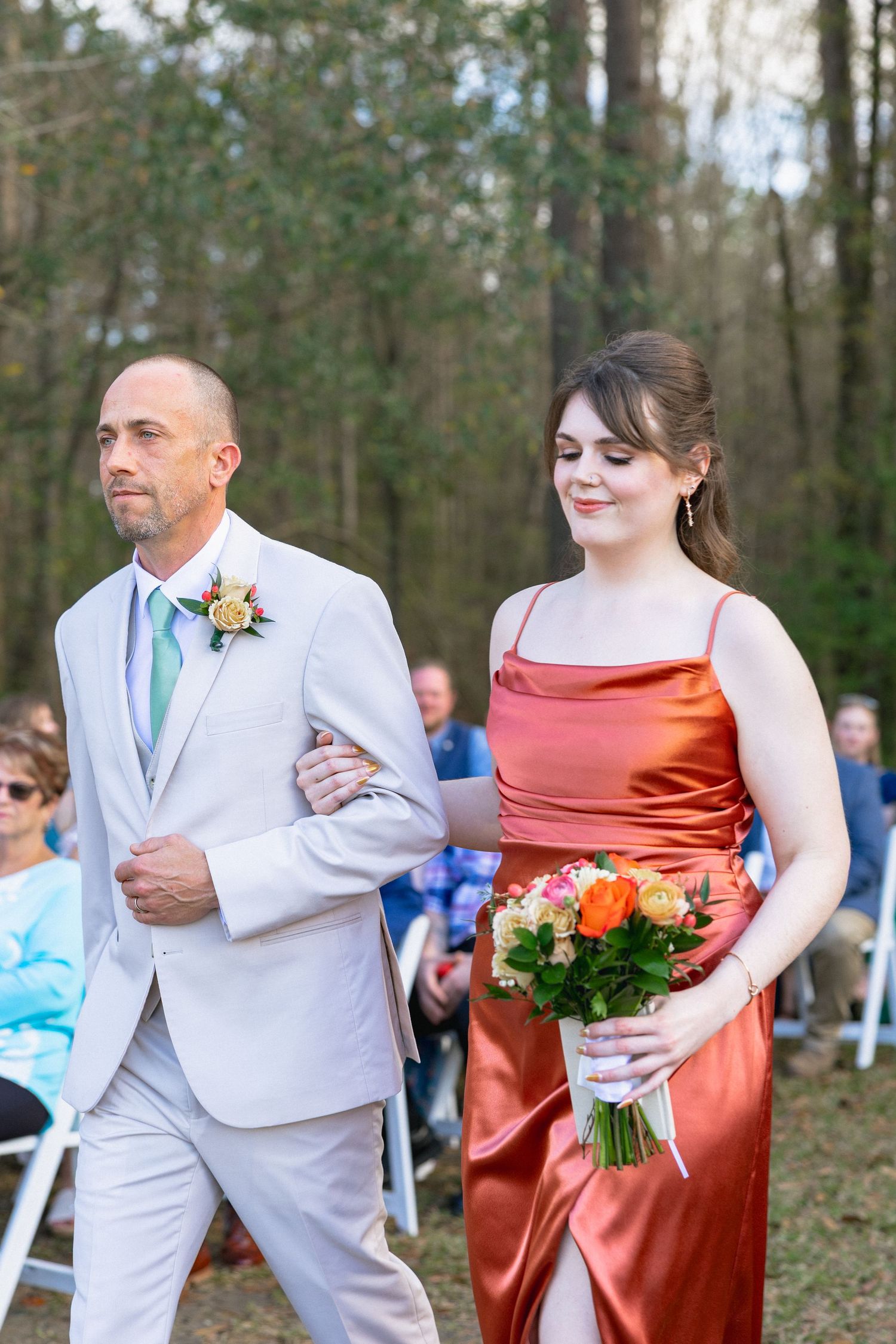 A groomsman in gray suit escorts a bridesmaid in rust satin gown down the outdoor wedding aisle while holding bouquet.