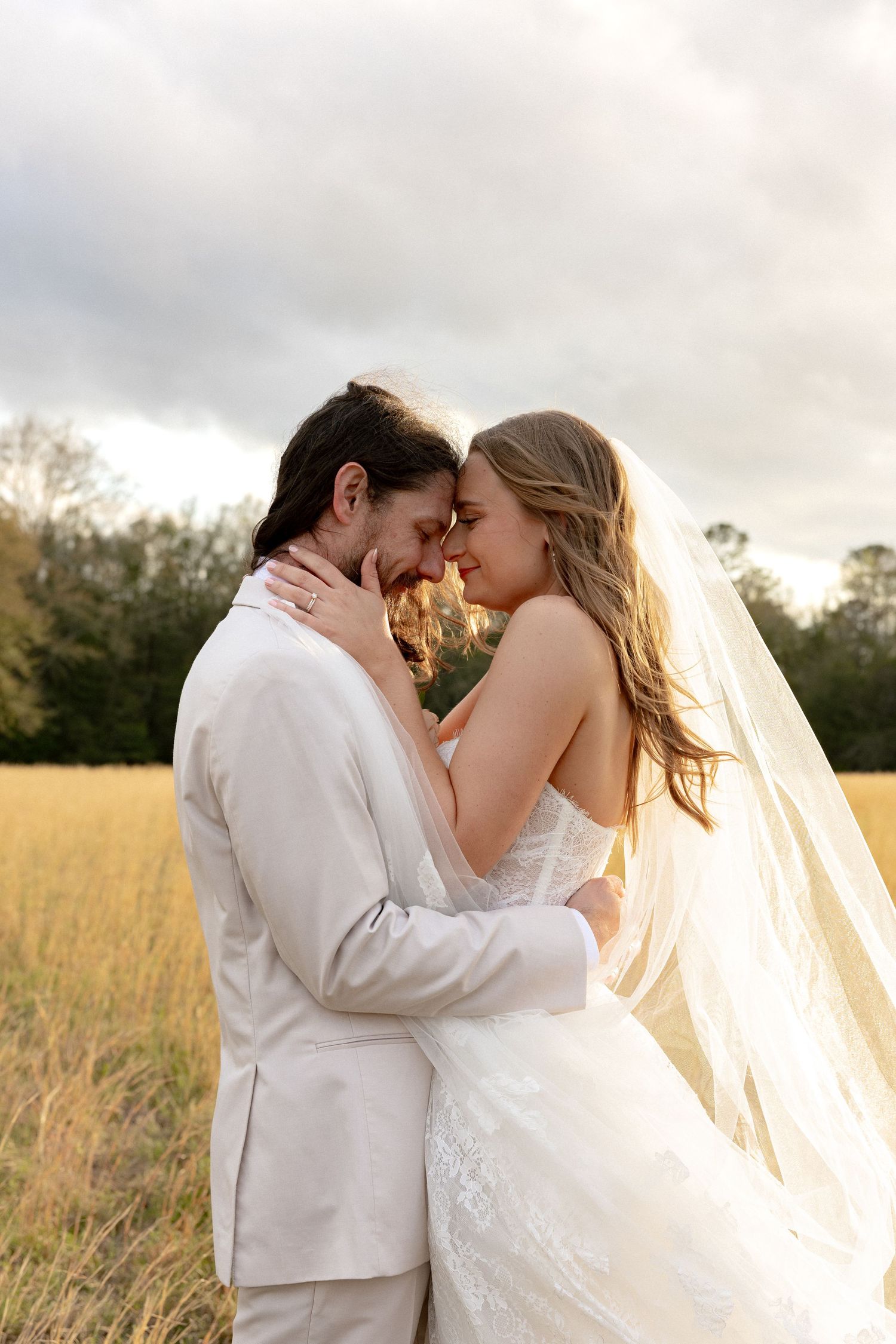 Romantic couple in white wedding attire touch foreheads in a sunlit golden meadow.
