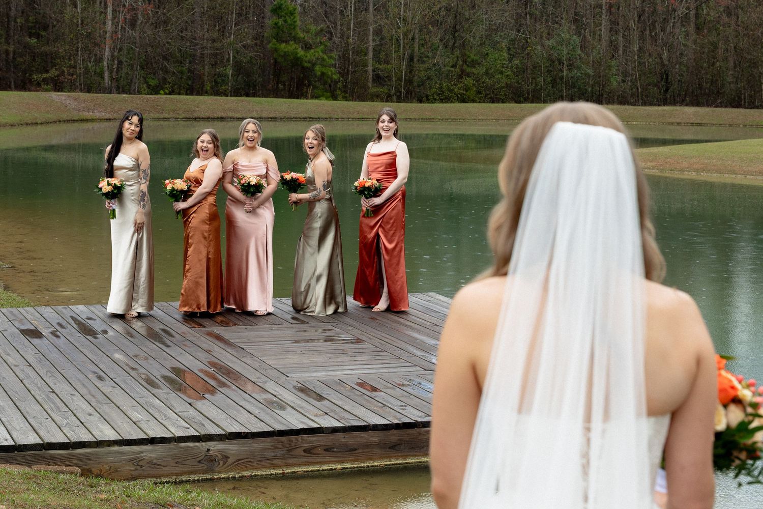 Bridesmaids in earth-tone satin dresses stand on wooden dock as bride approaches for first look.