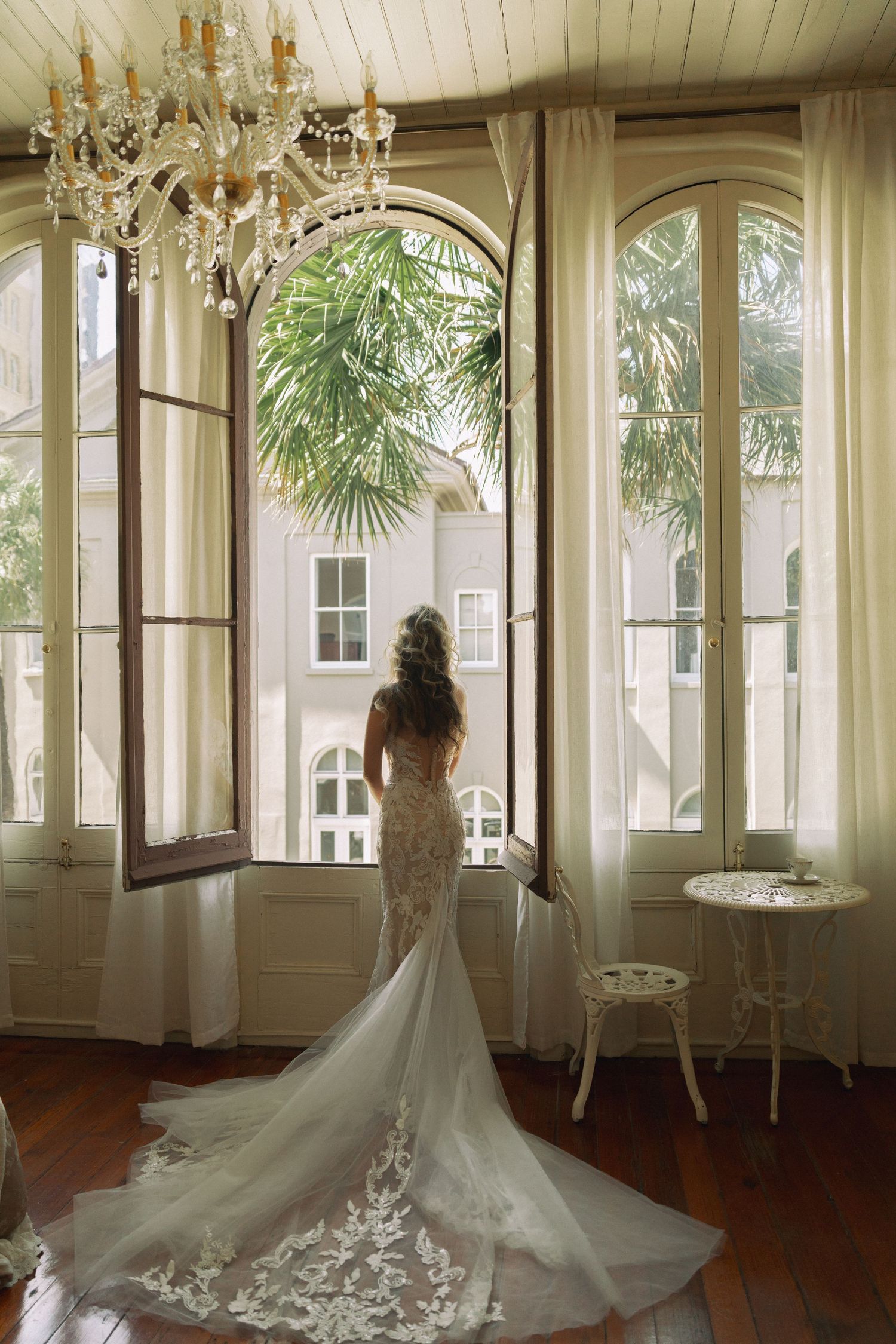 Bride in cathedral train lace gown stands at grand arched windows with chandelier in elegant room.