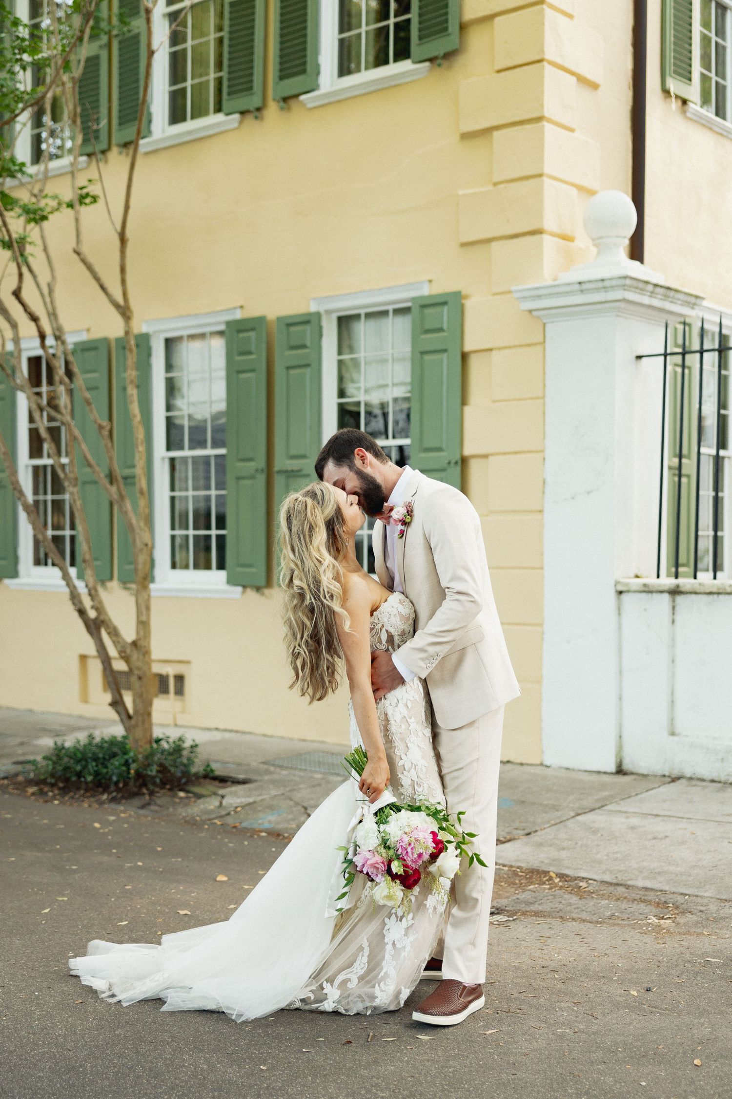 Bride and groom share a romantic kiss outside yellow Charleston historic building with green shutters and white pillar.